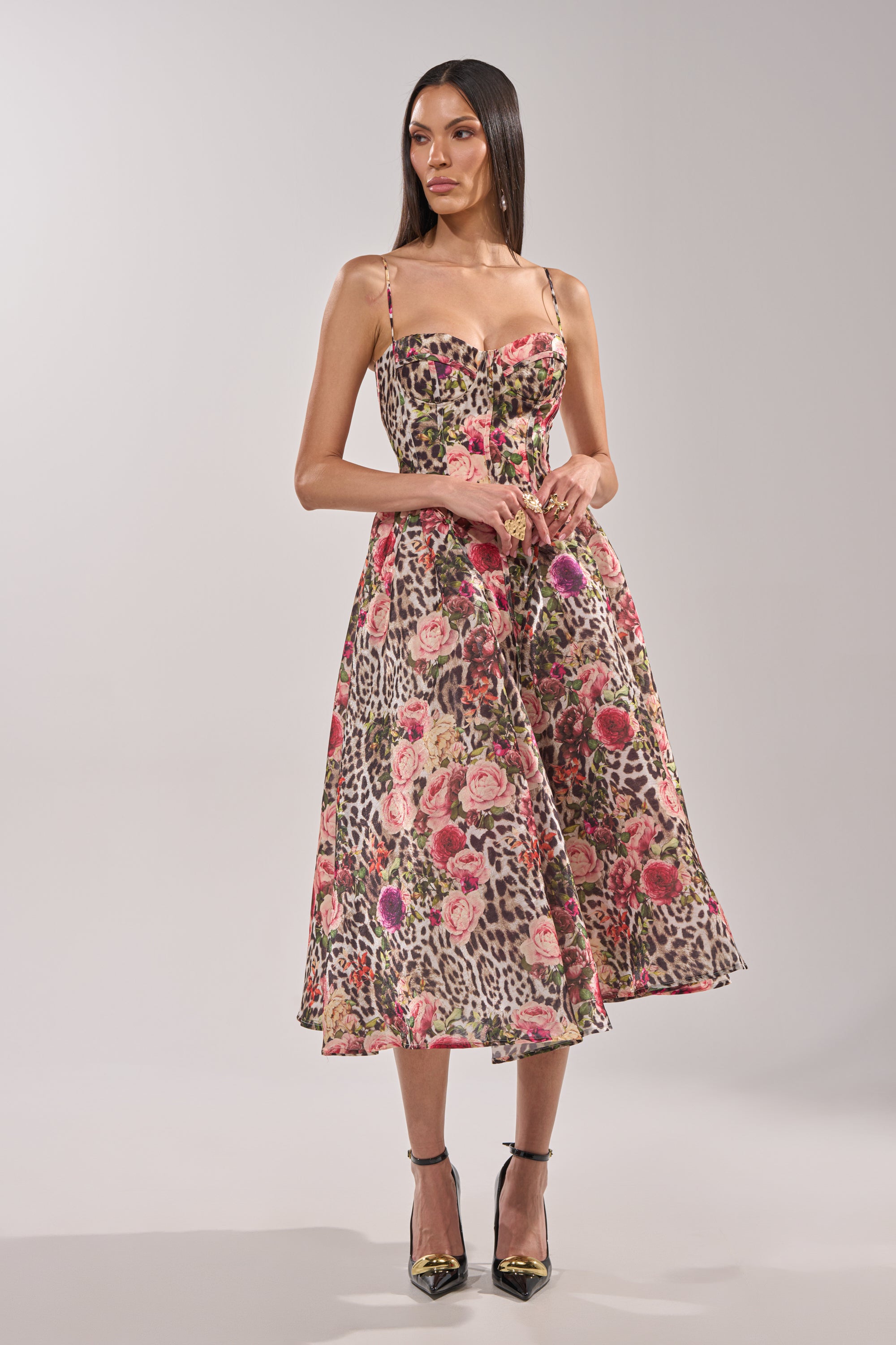 A woman with long, straight brown hair wears the COLETTE MAXI DRESS, a sleeveless piece with thin straps, corset bodice, and pink floral-leopard print. She completes the look with black ankle-strap heels. 1