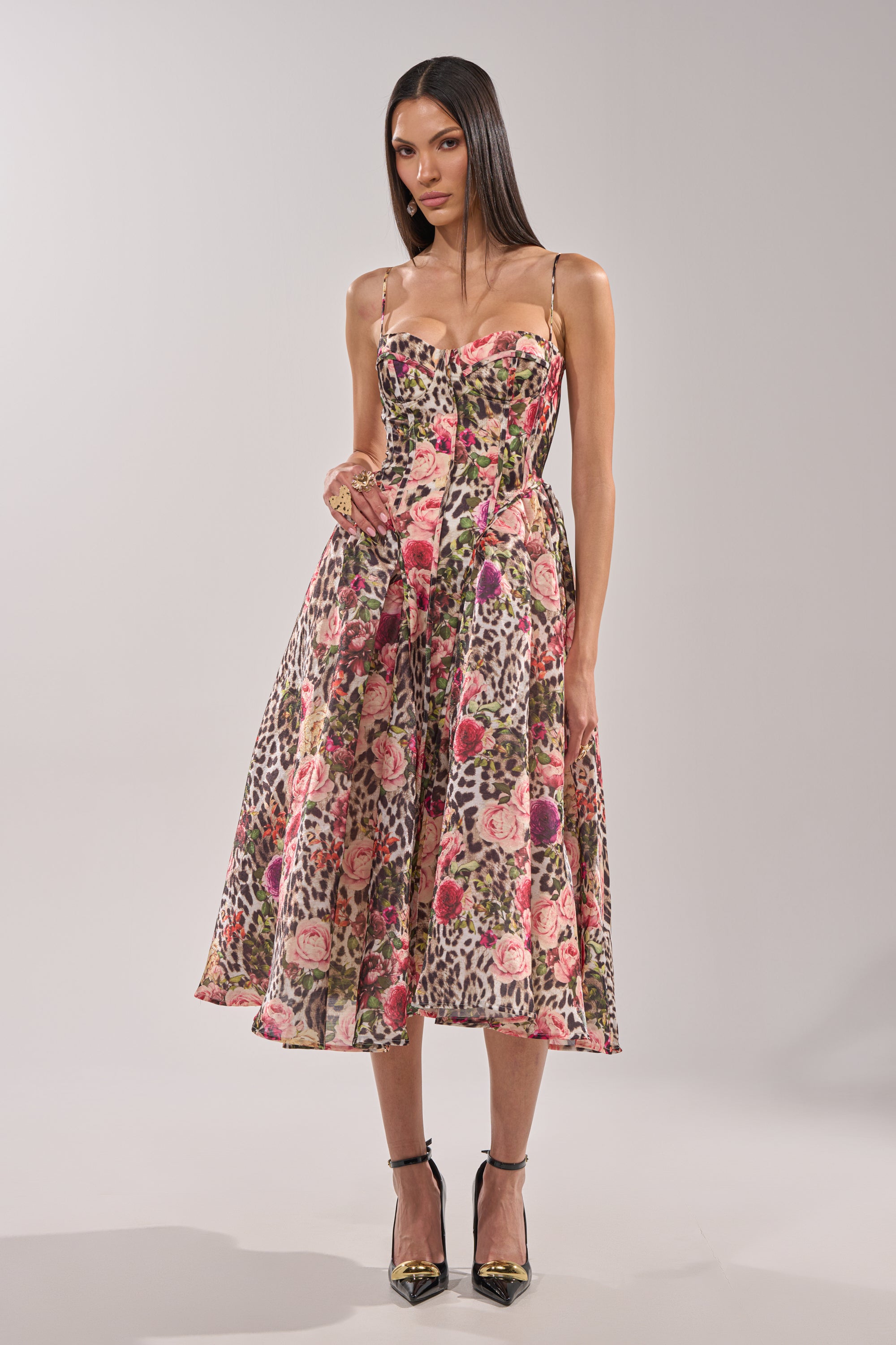 A woman wears the COLETTE MAXI DRESS, a sleeveless, flared dress with a corset bodice, featuring vibrant pink florals and leopard print, styled with black ankle-strap heels. 2