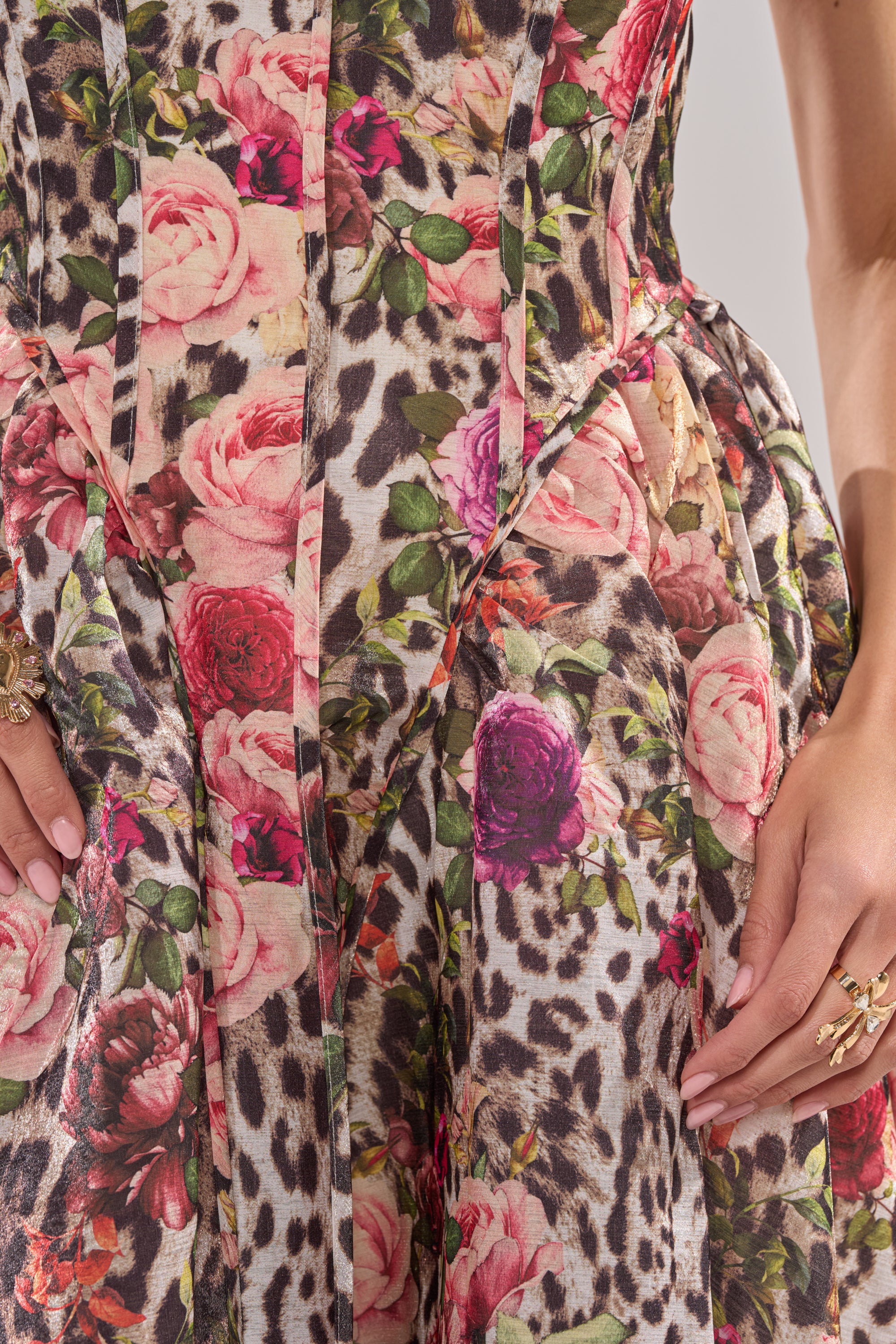Close-up of the COLETTE MAXI DRESS, showcasing a striking leopard print mixed with colorful roses and green leaves. A woman's hands adorned with gold rings rest at her sides, set against the textured fabric. 5