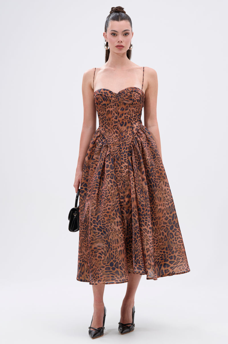 A woman models the CHEETAH SISTERS MIDI DRESS with a brown and black leopard print, thin straps, corset-style bodice, and flared skirt. She pairs it with a small black handbag and black heels against a plain light background.