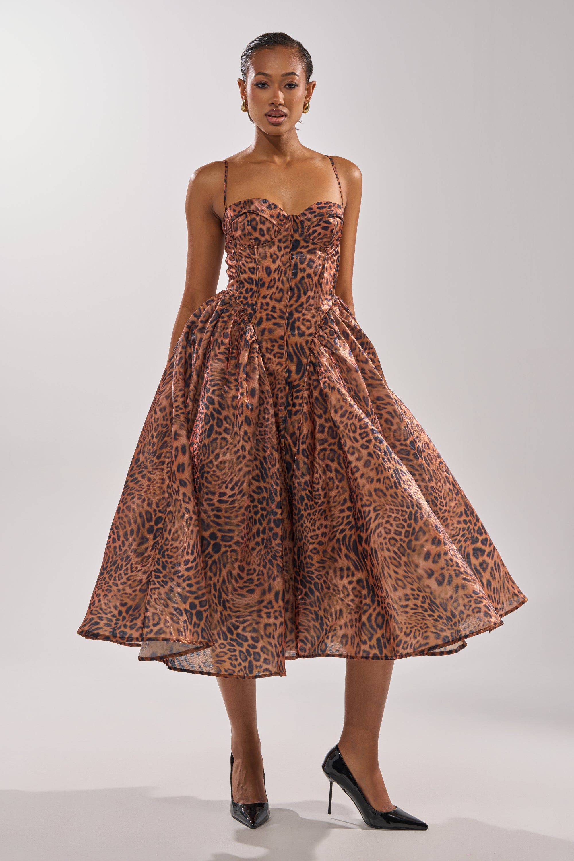 A woman models the CHEETAH SISTERS MIDI DRESS, a fit and flare midi with bold cheetah print, thin straps, and a full skirt, paired with black heels against a plain light backdrop.