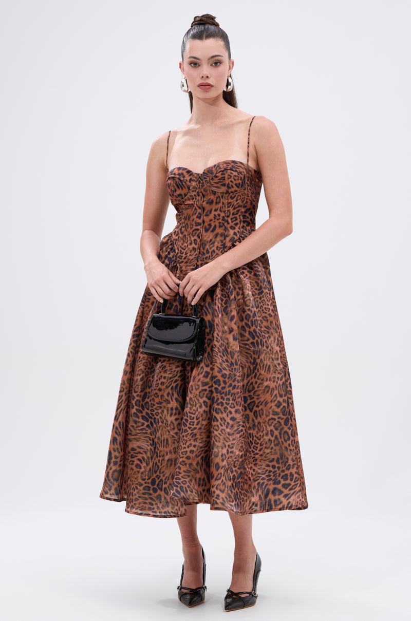 A woman in the CHEETAH SISTERS MIDI DRESS with a corseted bodice, black heels, and a small black handbag stands against a plain light background.