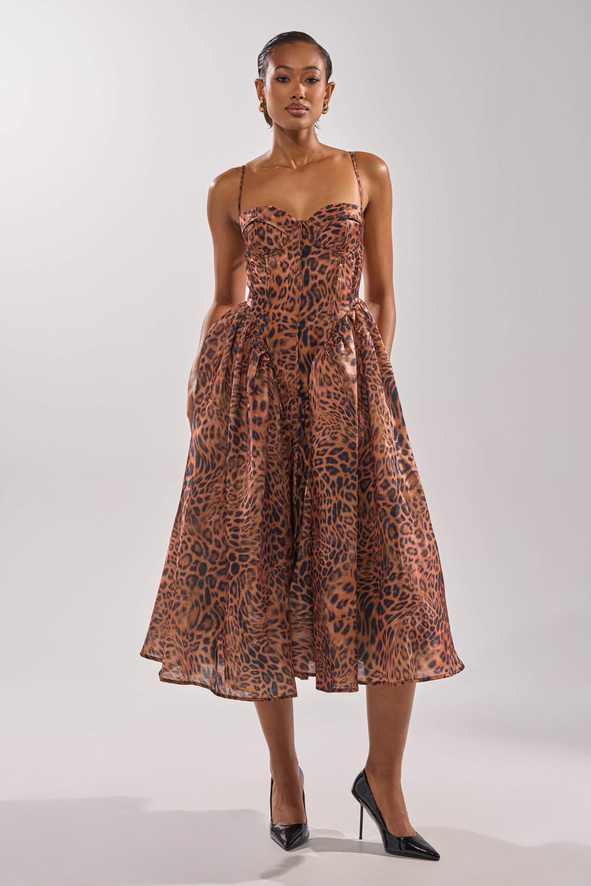 A woman stands in a studio wearing the CHEETAH SISTERS MIDI DRESS, featuring a brown leopard print, fitted corset bodice, and flared midi skirt. Her hair is pulled back and she pairs the look with black pointed-toe heels.