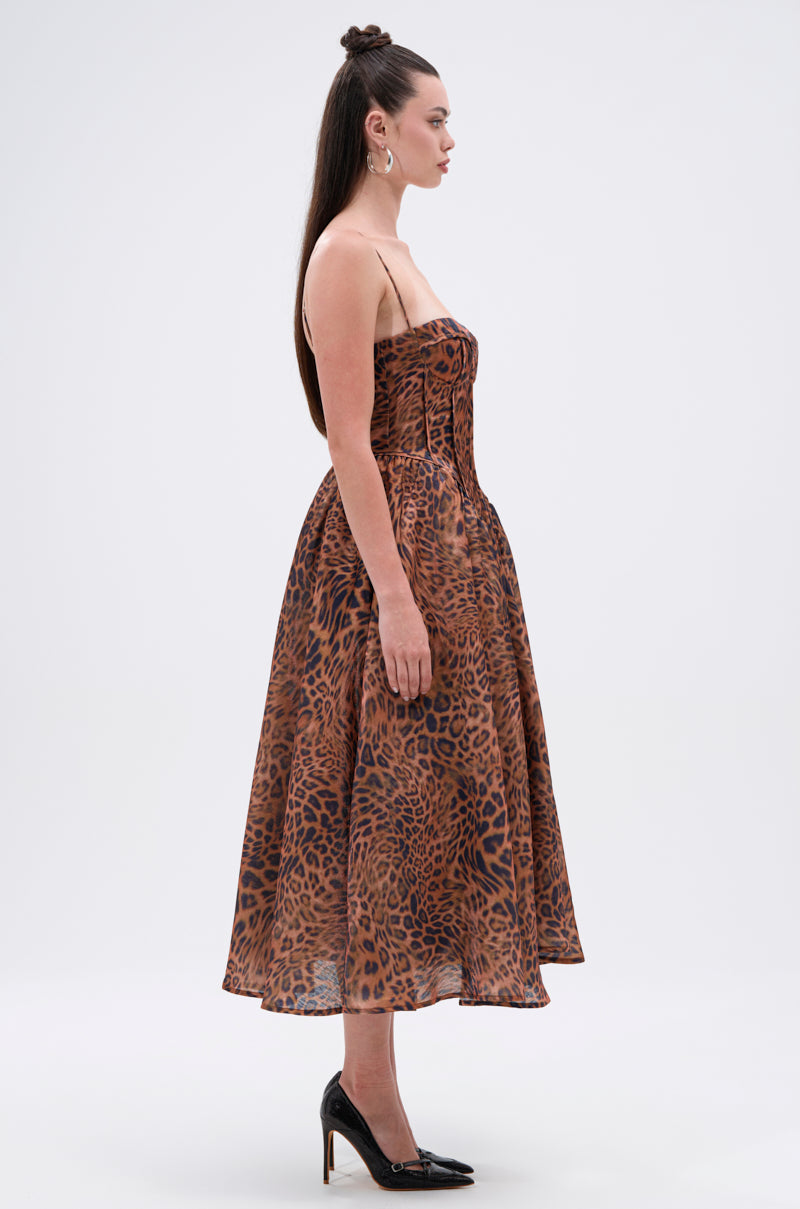 A woman in profile wears the CHEETAH SISTERS MIDI DRESS—a sleeveless, calf-length dress with thin straps and bold leopard print—paired with black high heels. Her long, straight hair flows against a plain background.
