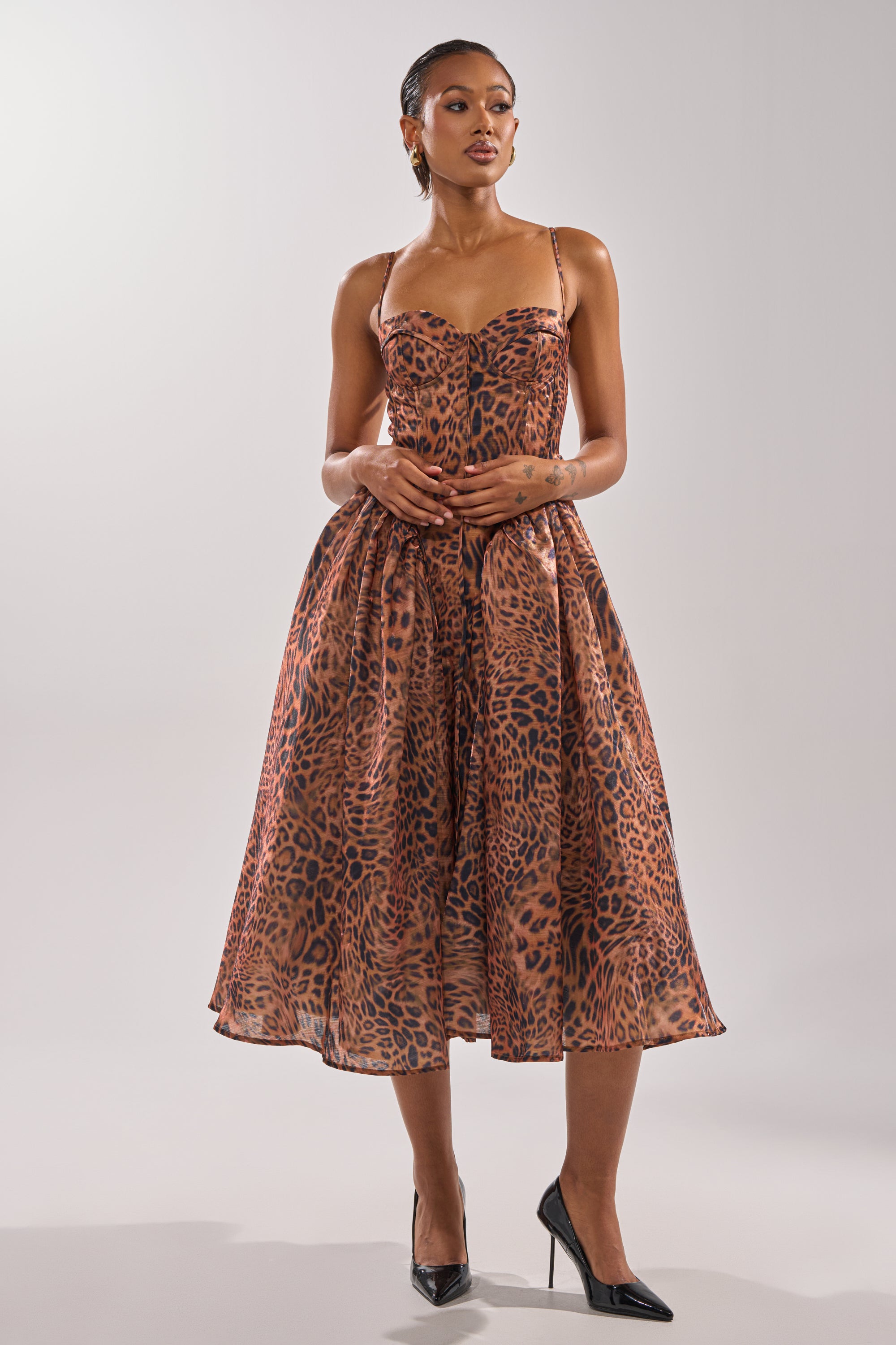 A woman poses in a studio against a white background wearing the CHEETAH SISTERS MIDI DRESS, a sleeveless, fitted midi dress with a corseted bodice and bold brown and black cheetah print. She completes the look with slicked-back hair and black pointed-toe heels.