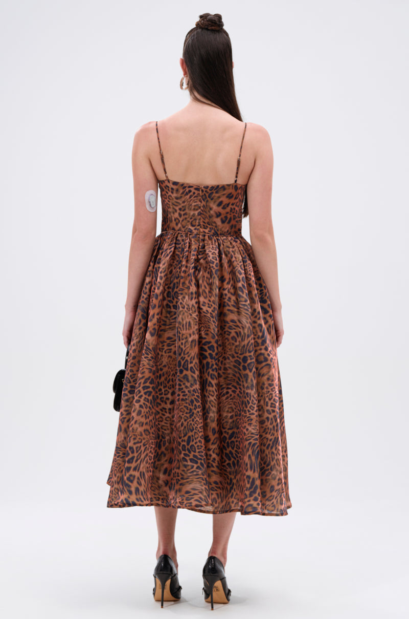 A woman faces away, wearing the CHEETAH SISTERS MIDI DRESS with a corseted bodice and brown and black leopard print. She holds a small black bag, pairs it with black shoes, and styles her hair in a half-up bun.