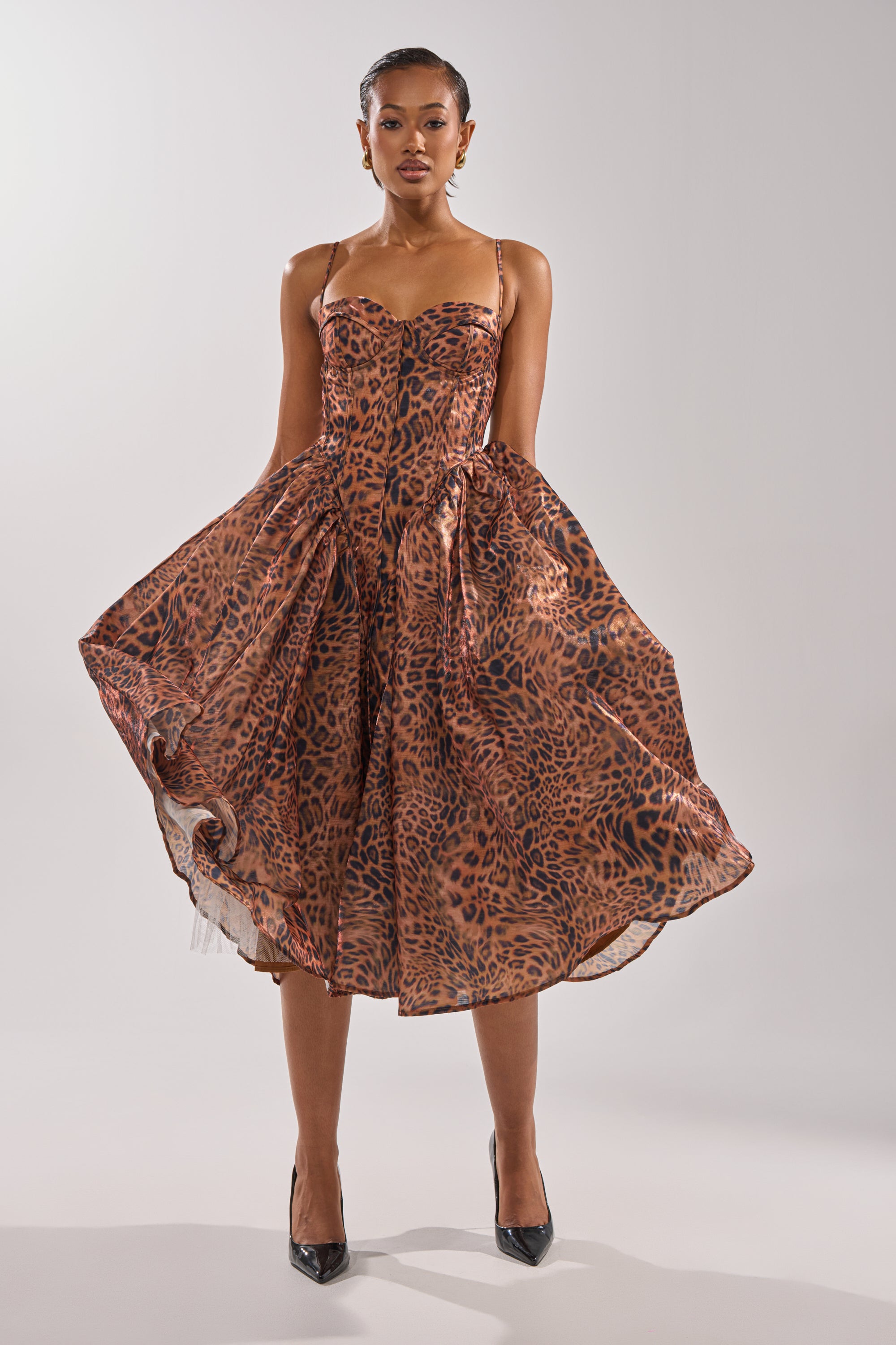 A woman stands confidently in the CHEETAH SISTERS MIDI DRESS, showcasing its corseted bodice and flared sleeveless leopard-print style, paired with black pointed-toe heels against a plain light background.
