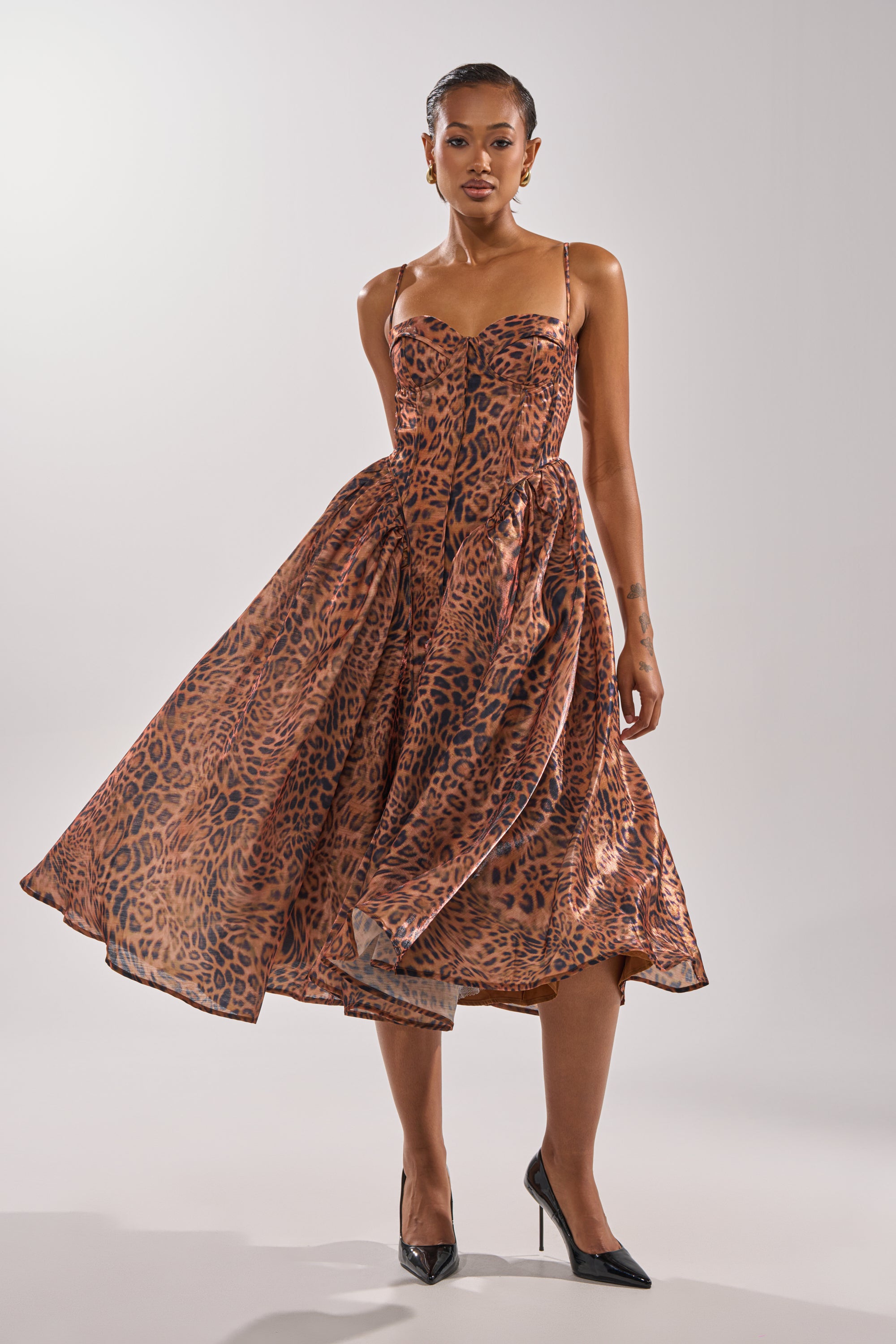 A woman wearing the CHEETAH SISTERS MIDI DRESS, a sleeveless fit and flare leopard print midi with thin straps and full skirt, stands in black pointed heels against a plain light background.