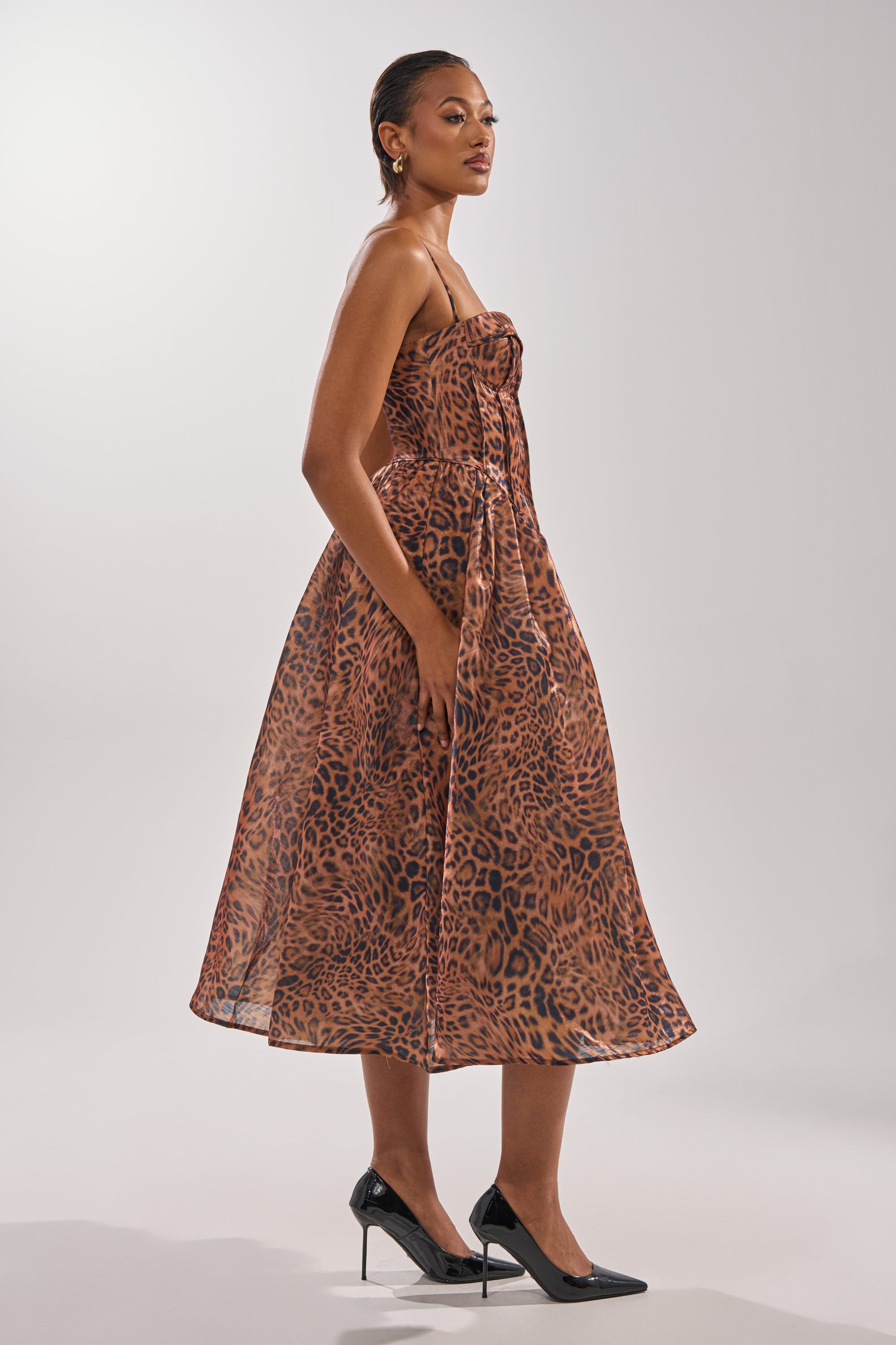 A woman in profile wears the CHEETAH SISTERS MIDI DRESS, a strapless, calf-length animal print design, paired with black high heels against a plain light background.