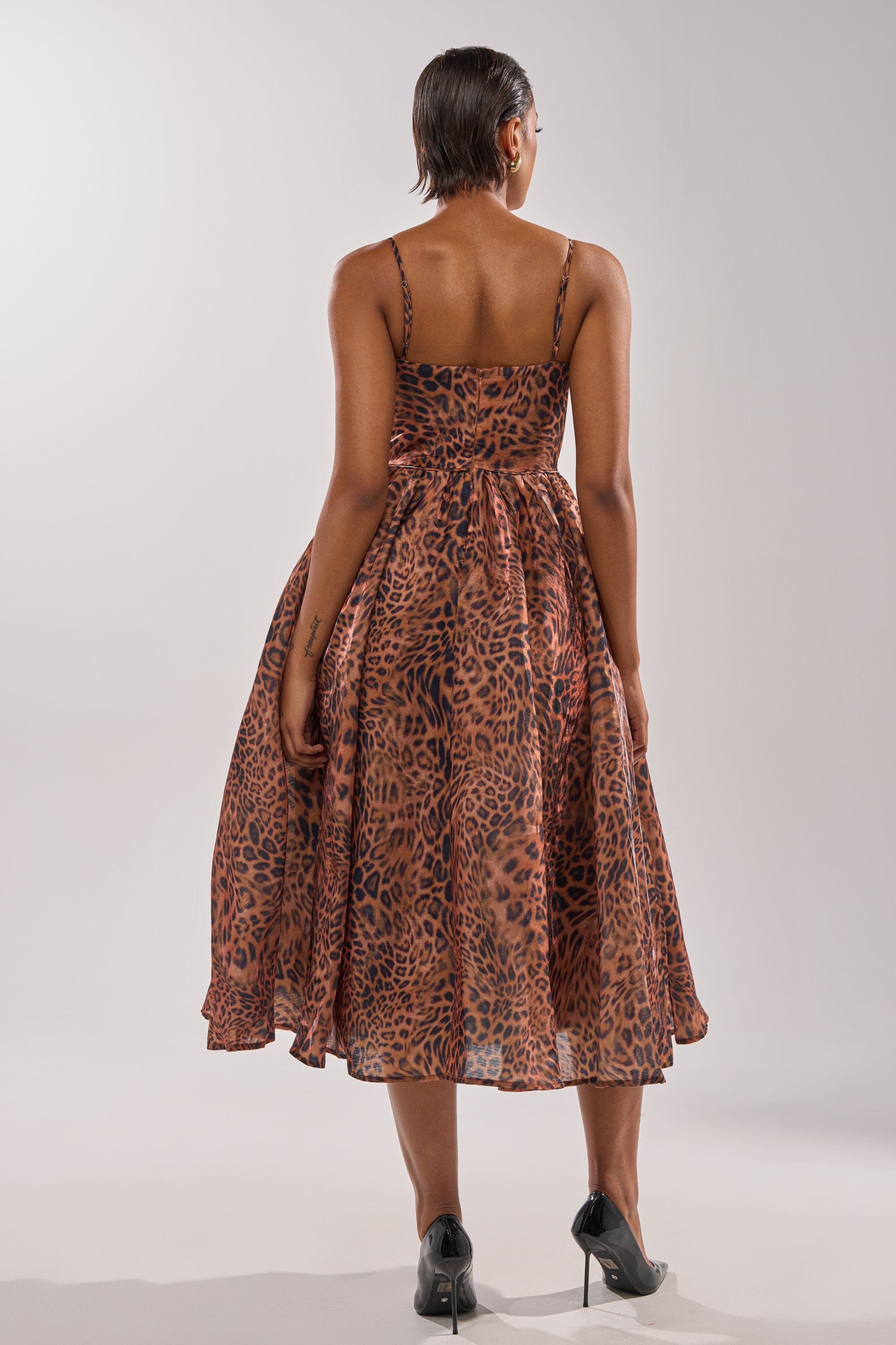 A person stands facing away, wearing the CHEETAH SISTERS MIDI DRESS—a sleeveless, leopard print midi dress with thin straps—paired with black high-heeled shoes against a plain light background.