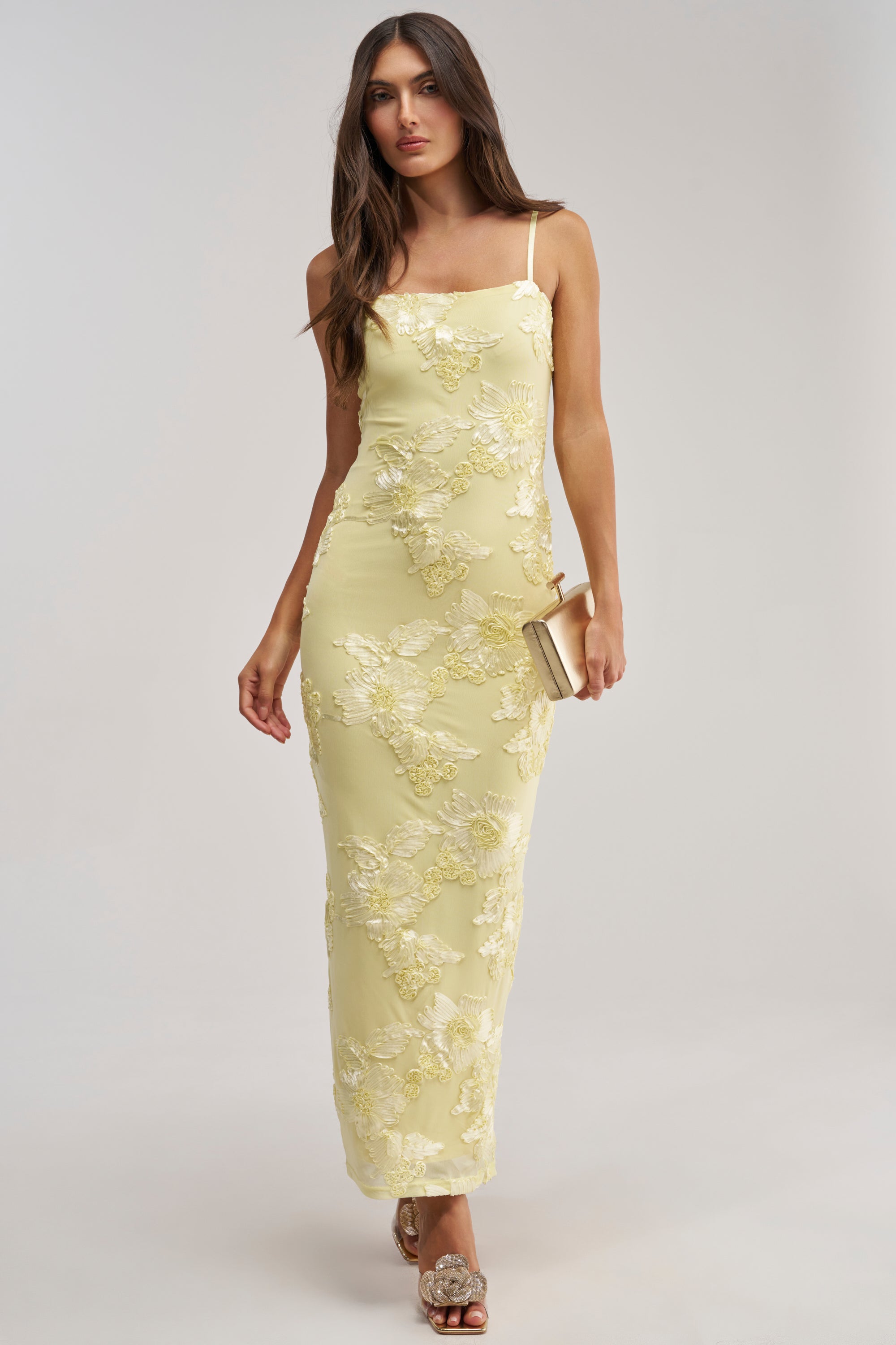 Wearing the TRY TO KEEP UP FLORAL MIDI DRESS IN YELLOW, a woman with loose brown hair holds a beige clutch and wears open-toe heels. This sleeveless, fitted floral piece is perfect for summer weddings.