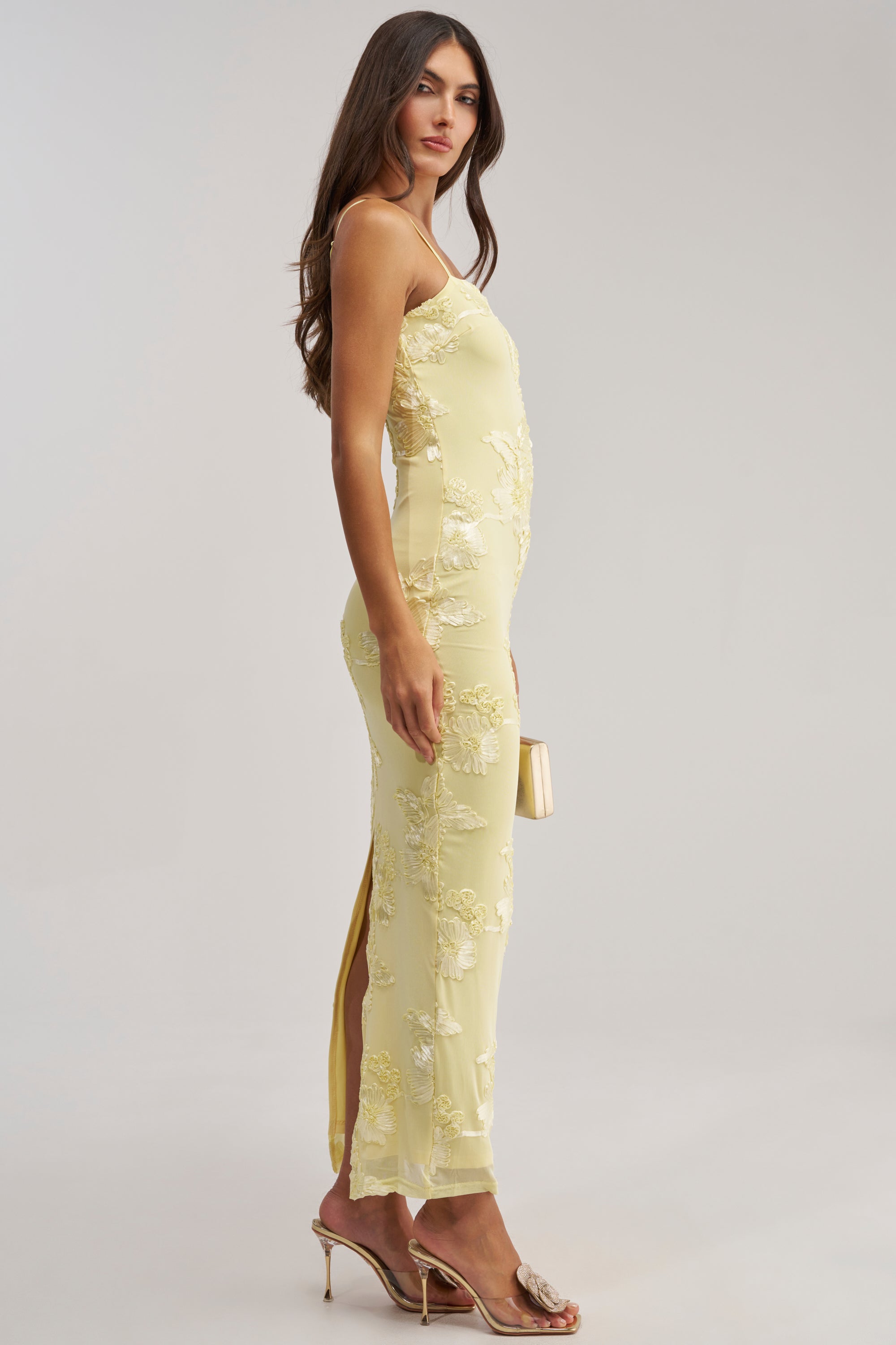 A woman stands sideways in the TRY TO KEEP UP FLORAL MIDI DRESS IN YELLOW, featuring elegant embroidery and a long back slit. She holds a beige clutch and wears transparent heels—an ideal chic wedding guest dress against a plain background.