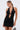 A woman stands against a light background, looking down while holding a purse and wearing the DANCING QUEEN BEADED MINI DRESS, featuring black sequins, a halter neckline, cutouts, and a deep V front.