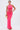 A woman stands against a white background wearing the BRIGHT AS THE SKY MESH MAXI DRESS, a sleeveless hot pink maxi with halter straps and sheer midsection, paired with a beige clutch, hoop earrings, and red sandals.