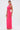 A woman wears the BRIGHT AS THE SKY MESH MAXI DRESS, a vibrant pink halter maxi with sheer side panels. She holds a beige clutch, pairs orange sandals, and styles her long straight hair in a low ponytail.