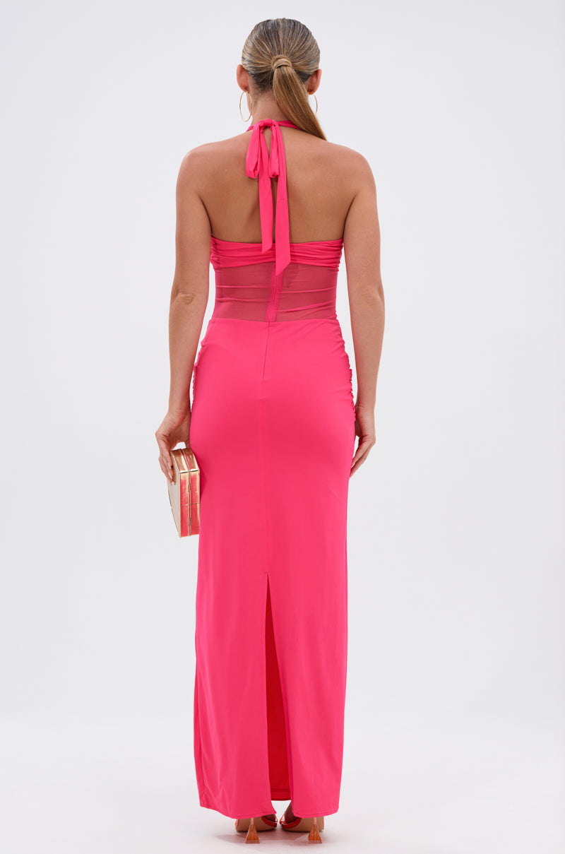 A woman with long hair in a low ponytail wears the BRIGHT AS THE SKY MESH MAXI DRESS—a bright pink halter dress with sheer panels and back slit—holding a gold clutch against a plain white background.