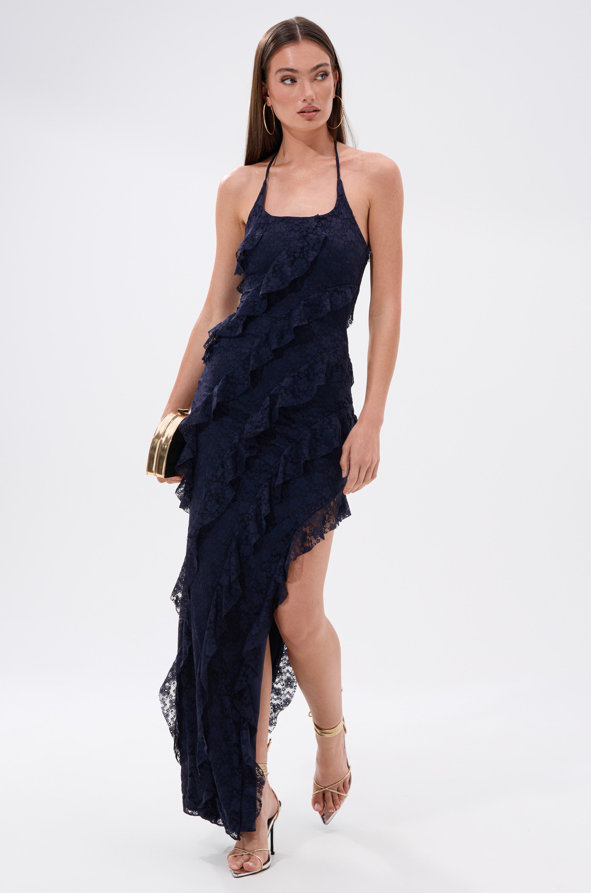 Wearing the "I GOT IT ALL LACE MIDI DRESS IN NAVY," a woman stands against a plain background, styling it with a gold clutch, gold hoop earrings, and strappy heels.