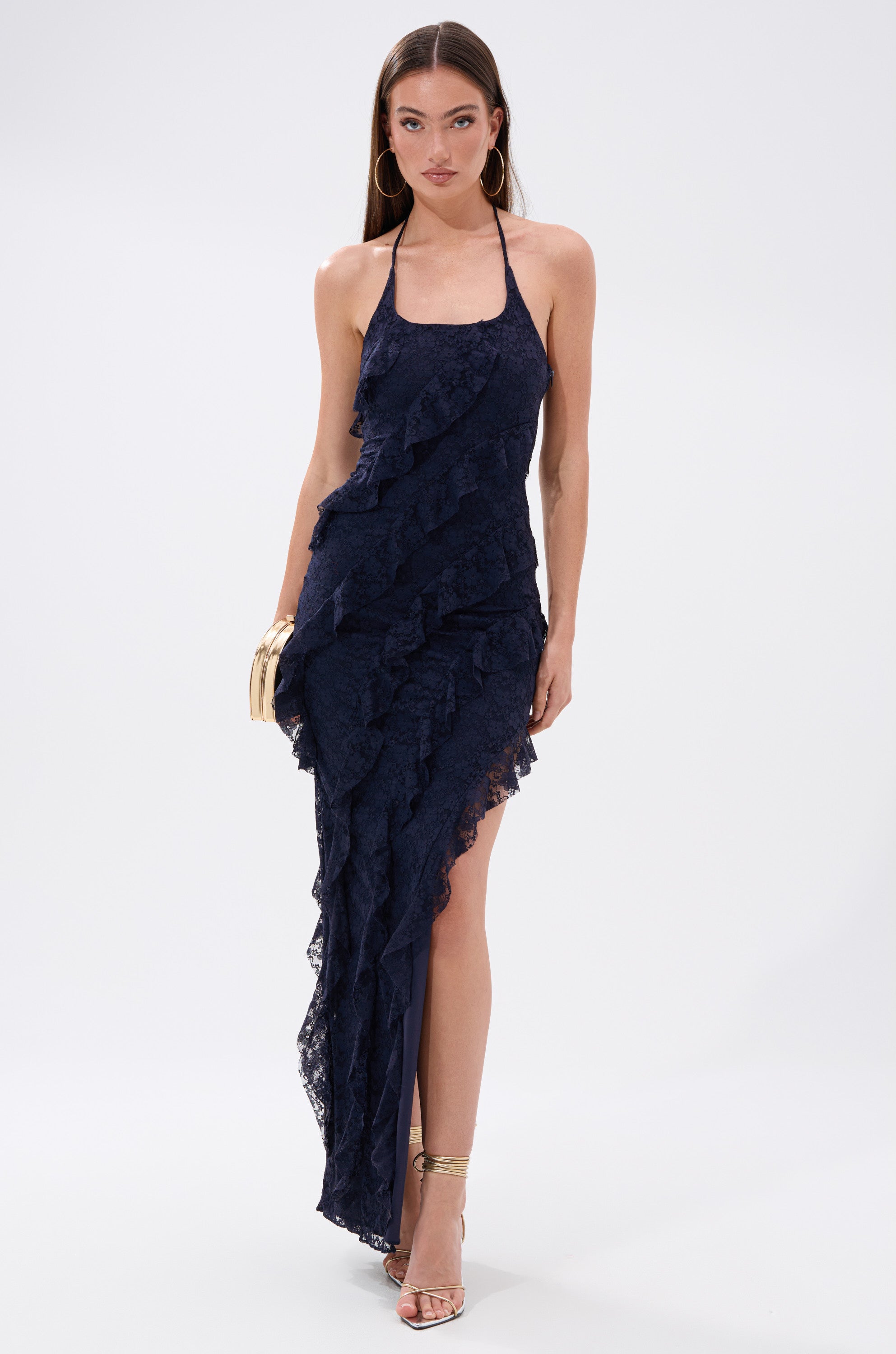 A woman stands against a white background wearing the I GOT IT ALL LACE MIDI DRESS IN NAVY, holding a small clutch and sporting strappy heels.