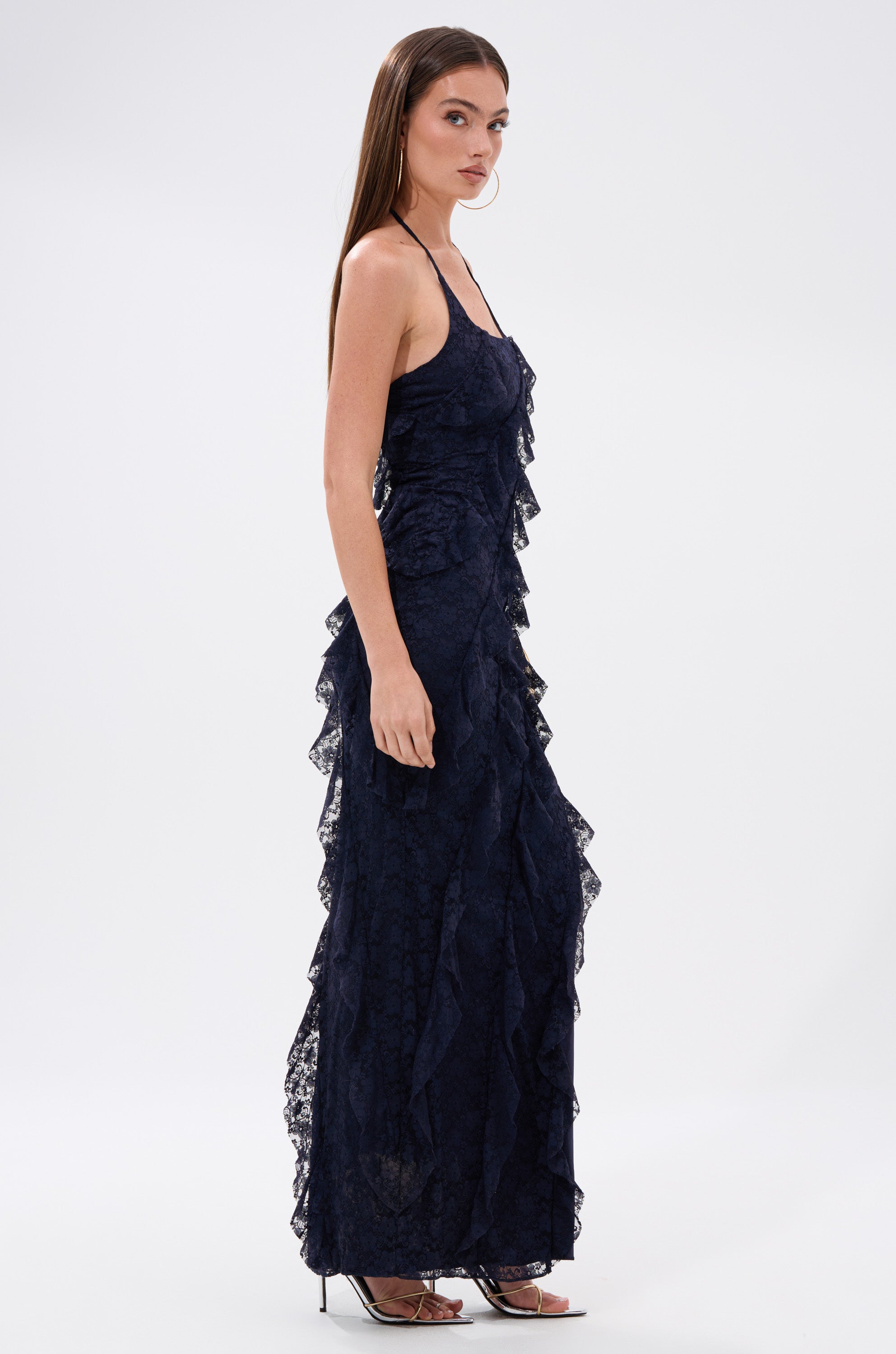 A woman models the I GOT IT ALL LACE MIDI DRESS IN NAVY, a sleeveless floral lace dress with ruffle details, paired with strappy heels and hoop earrings, standing sideways against a plain white background.