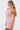 A woman with long, straight hair in a ponytail models the PEARL QUEEN MINI DRESS, a fitted pink party dress with spaghetti straps and delicate beads. She stands in profile against a plain white background, looking left.