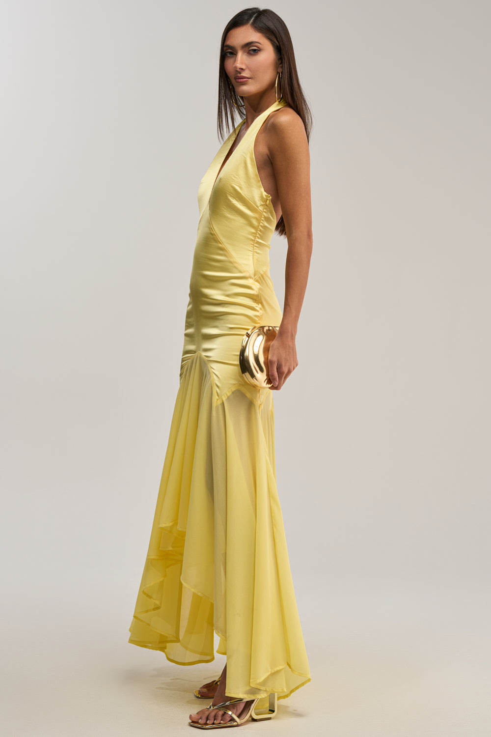 A woman in the BET YOU WON'T Satin Midi Dress in yellow stands sideways, holding a gold clutch. She wears gold hoop earrings, has long straight hair, and strappy yellow heels—an effortless wedding guest look against a light background.