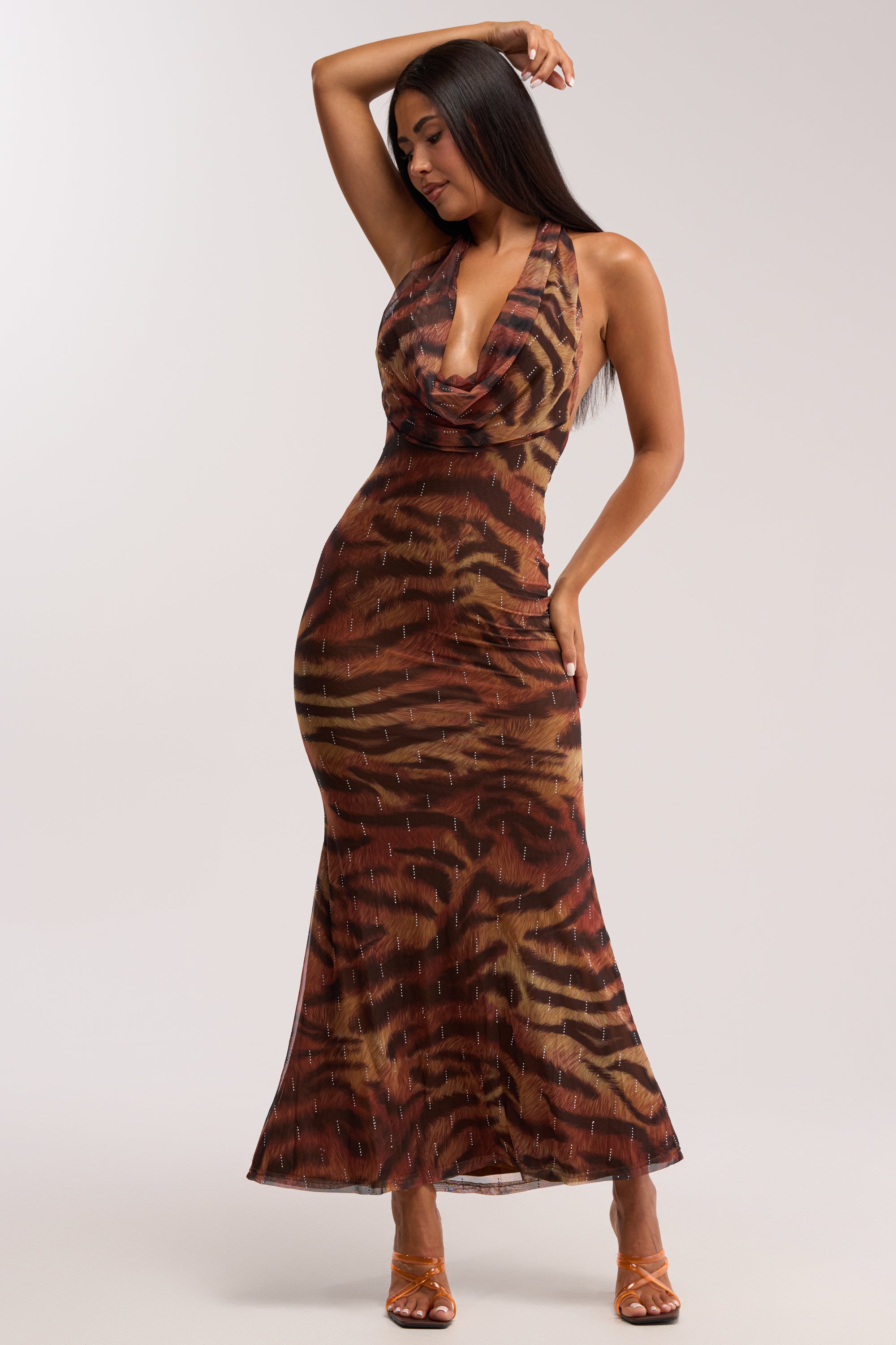 A woman models the PRESSURE STAYS ON CHEETAH MIDI DRESS, a sleeveless cowl neck halter style with a bold cheetah print, posing confidently with one hand on her hip and the other in her hair, paired with open-toe heels.