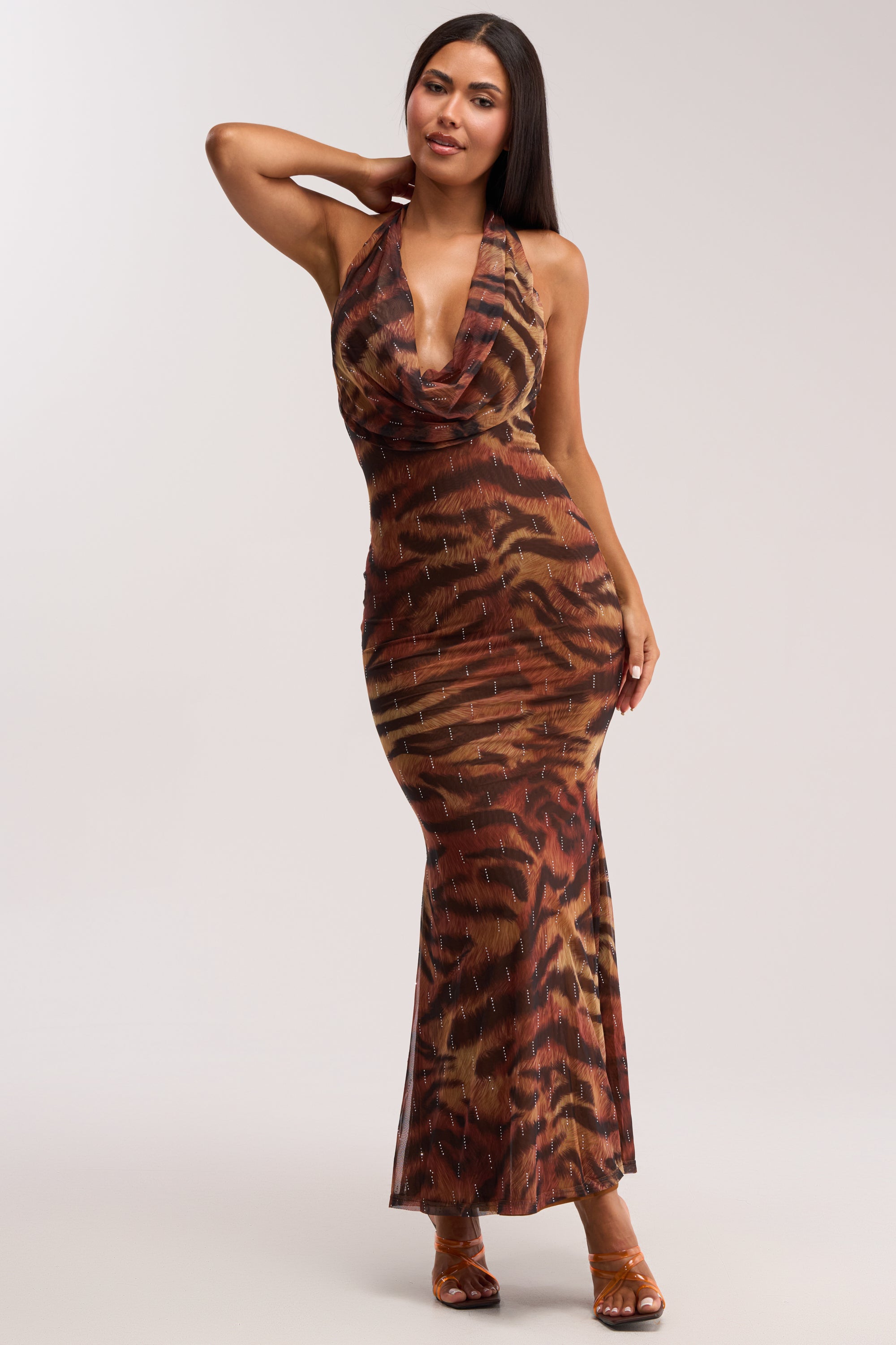 A woman poses in the PRESSURE STAYS ON CHEETAH MIDI DRESS, a sleeveless cowl neck halter dress with a brown and orange animal print, deep neckline, sparkling accents, and brown open-toe heels—an effortlessly sexy summer look.