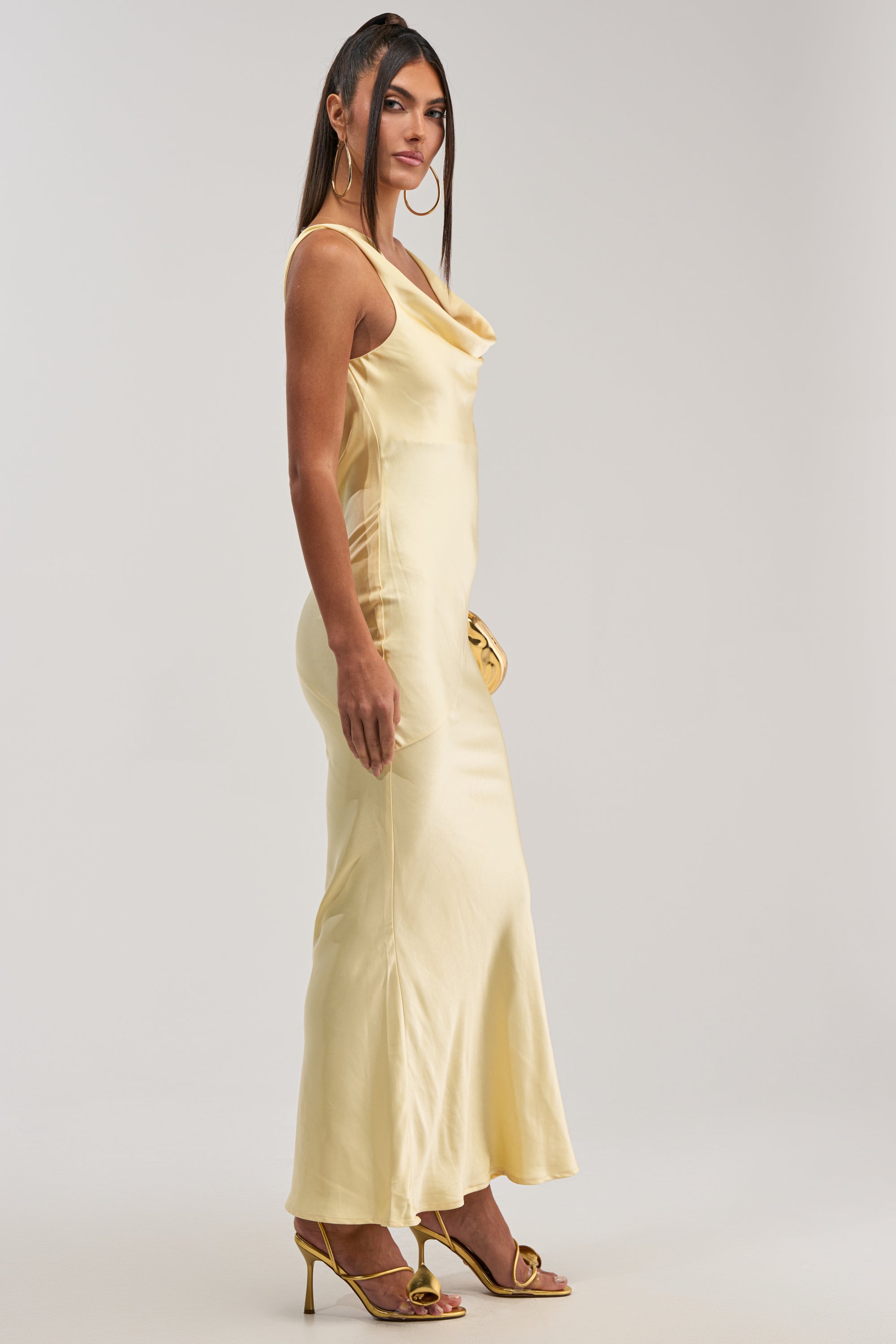 A woman in profile models the SOFT LAUNCH SATIN MAXI DRESS IN BUTTER YELLOW with gold hoop earrings, high-heeled sandals, and a yellow clutch—the perfect wedding guest look—against a plain light background.