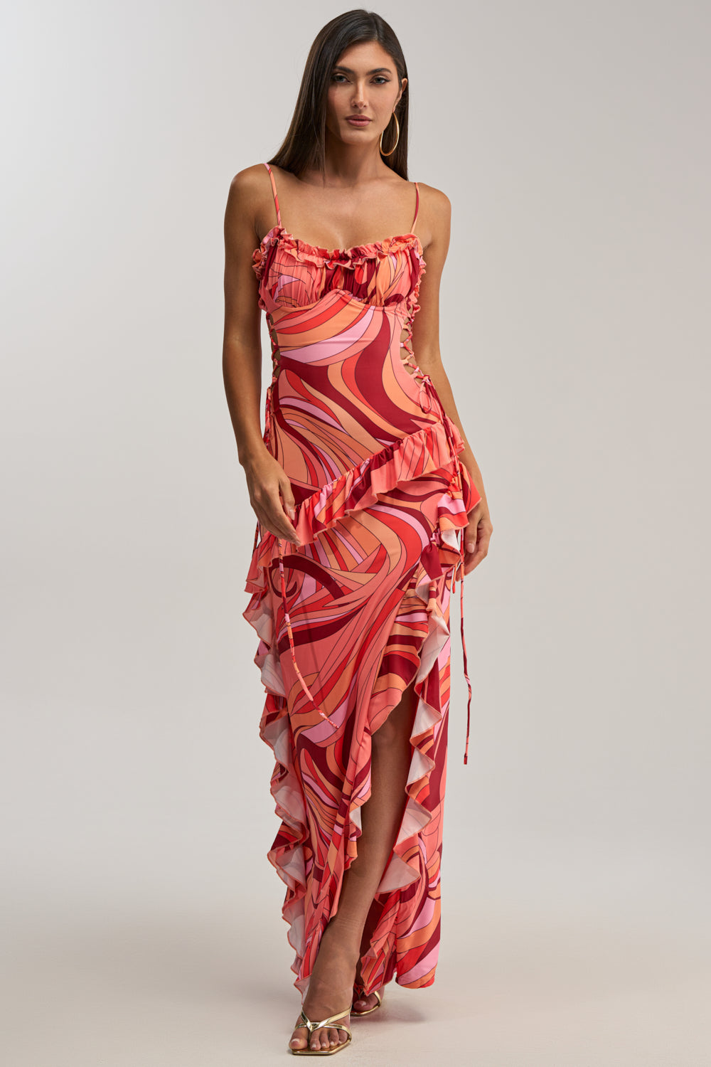 A woman stands in high-heeled sandals against a plain background, modeling the HANDLE WITH CARE ABSTRACT MIDI DRESS IN ORANGE—a sleeveless, fitted dress with pink and red swirls, ruffle details, and an asymmetrical hem—perfect for wedding guests.