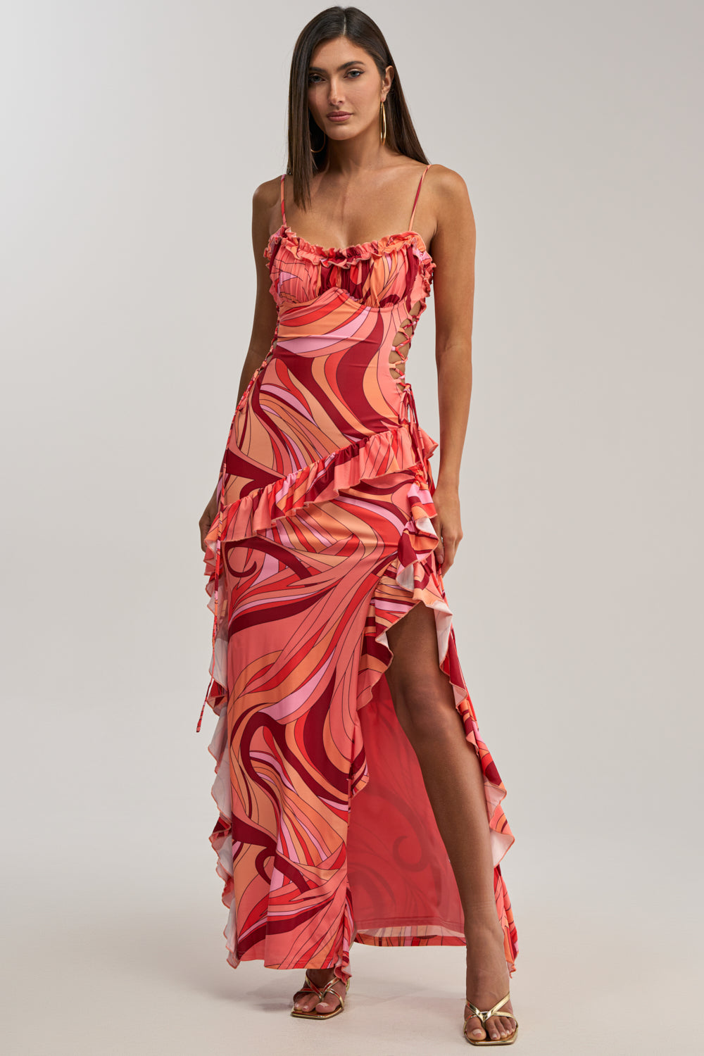 A woman wears the HANDLE WITH CARE ABSTRACT MIDI DRESS IN ORANGE, featuring red, pink, and peach swirling patterns, ruffle accents, a high side slit, paired with gold high-heeled sandals.