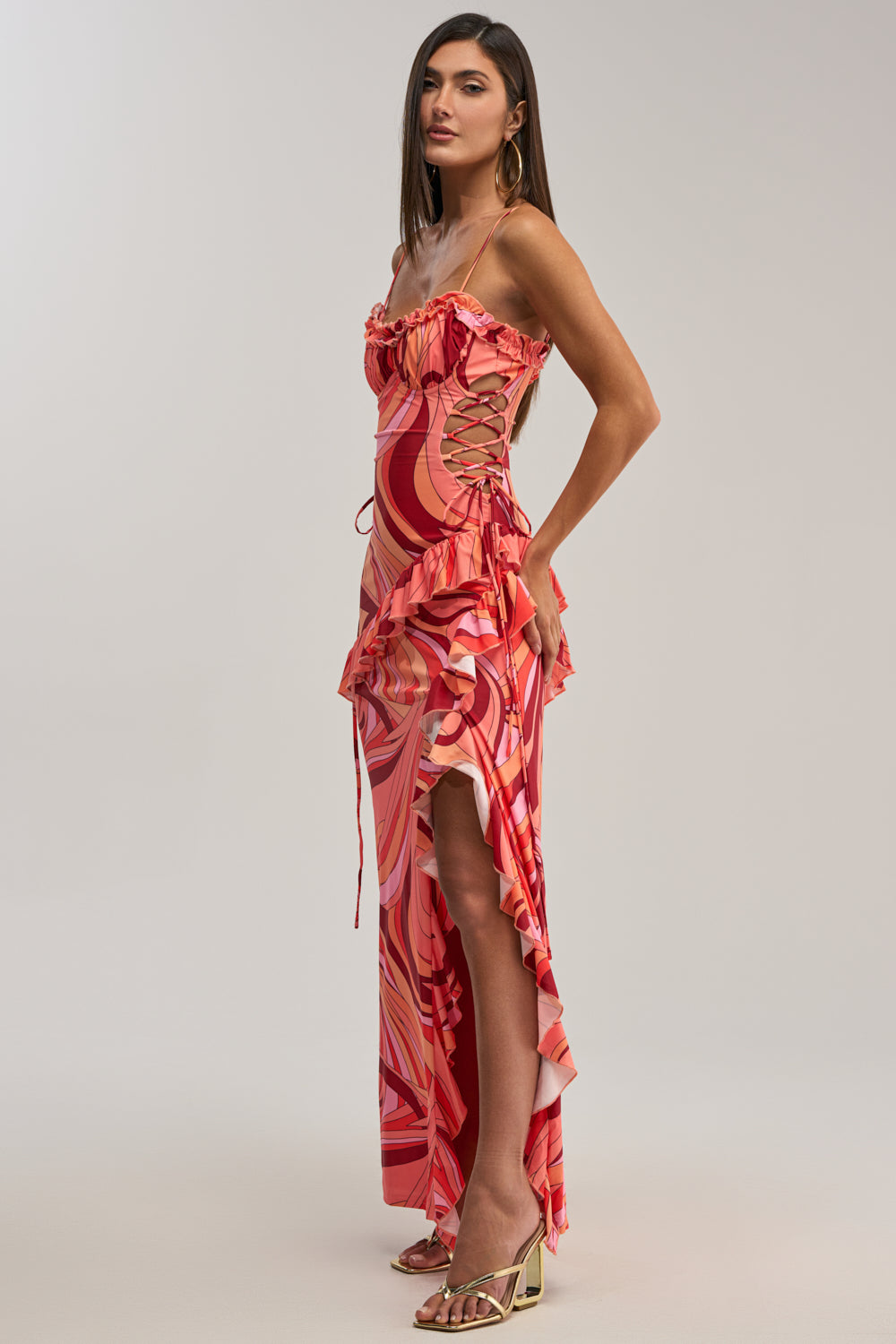 Woman in profile wears the HANDLE WITH CARE ABSTRACT MIDI DRESS IN ORANGE, a vibrant formal dress with ruffled details, side lace-up accents, and a high slit—ideal for wedding guests—paired with gold strappy heels against a plain background.