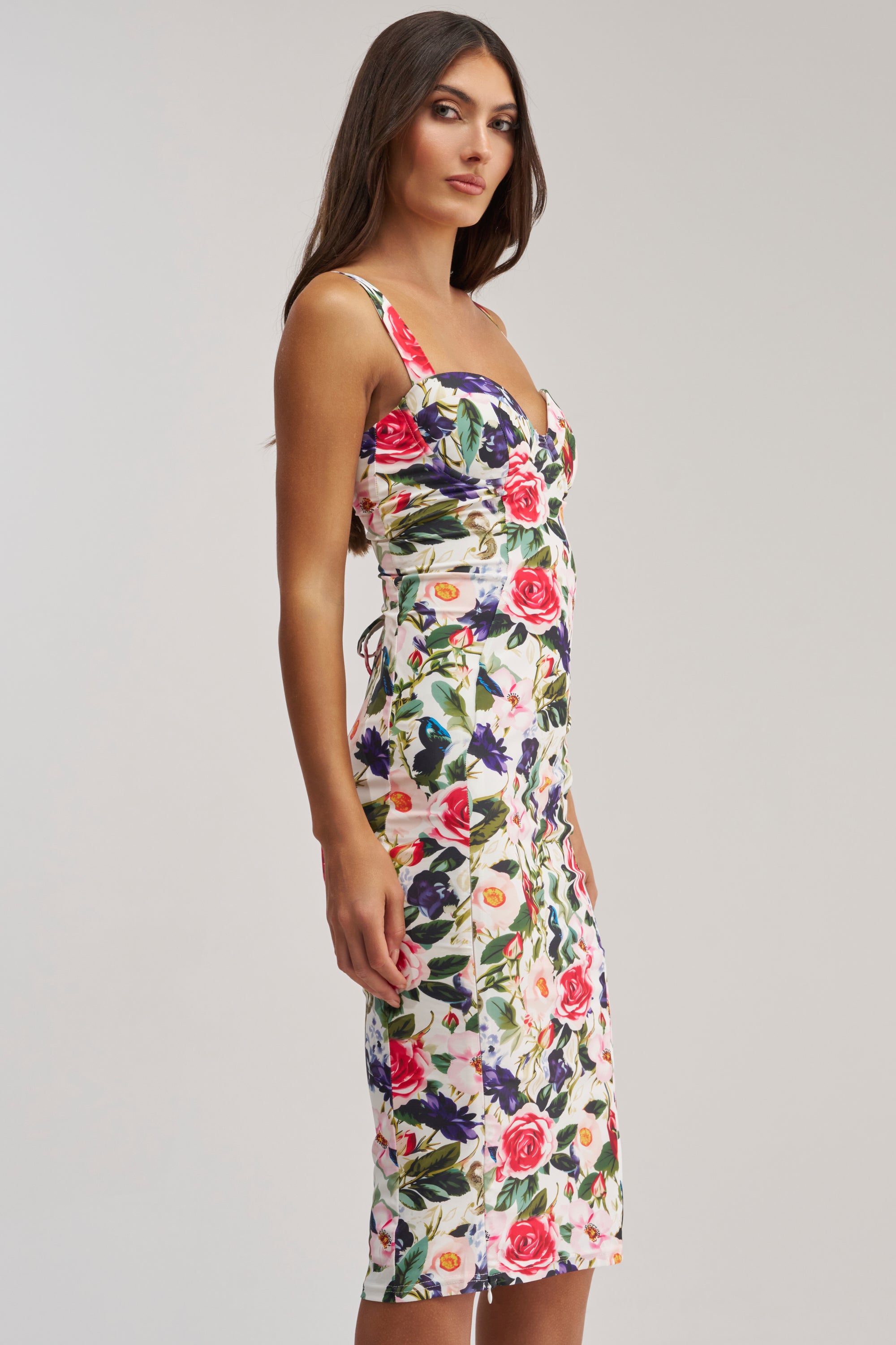 Model wears the TALK IS CHEAP FLORAL MIDI DRESS—a white bodycon midi with a floral print, sweetheart neckline, and lace-up corset back—standing against a plain background for a chic summer cocktail look.
