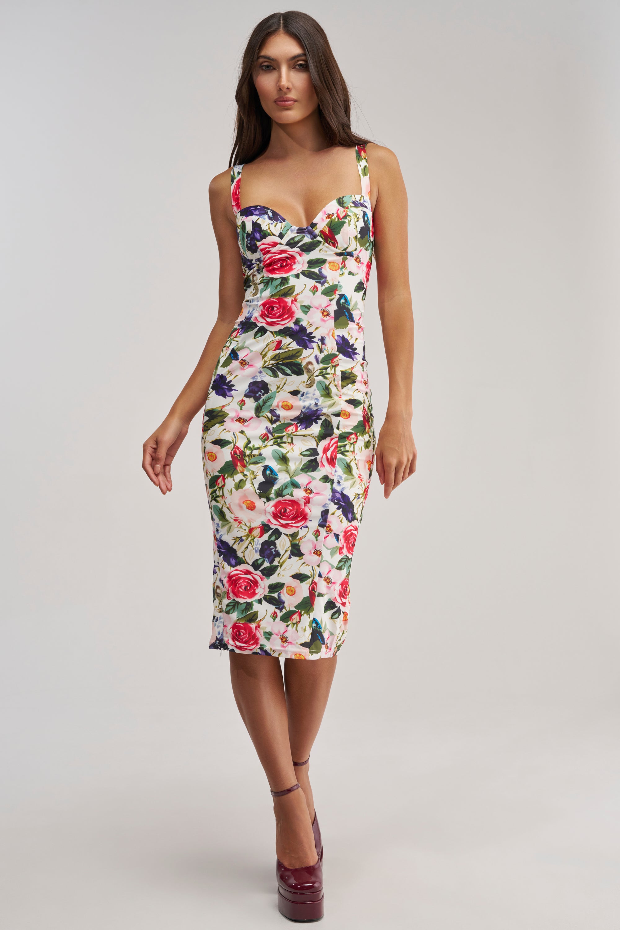 A woman in the TALK IS CHEAP FLORAL MIDI DRESS—white, floral, bodycon midi with a sweetheart neckline and lace-up corset back—pairs it with maroon platform heels for an elegant summer cocktail look against a light background.