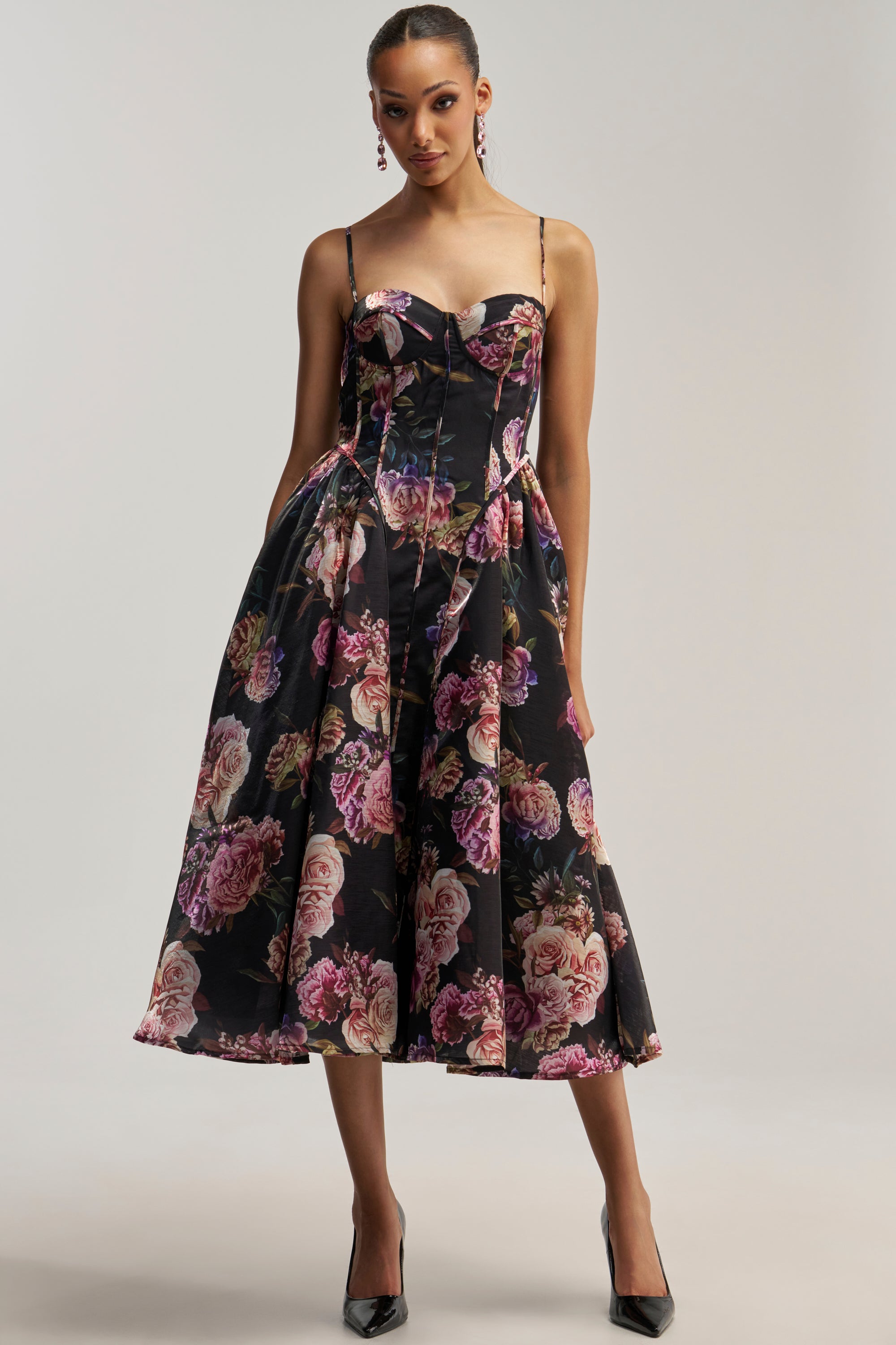A woman wearing the FEELING FLORAL MAXI DRESS IN PINK MULTI with spaghetti straps and black high heels stands against a plain light background.