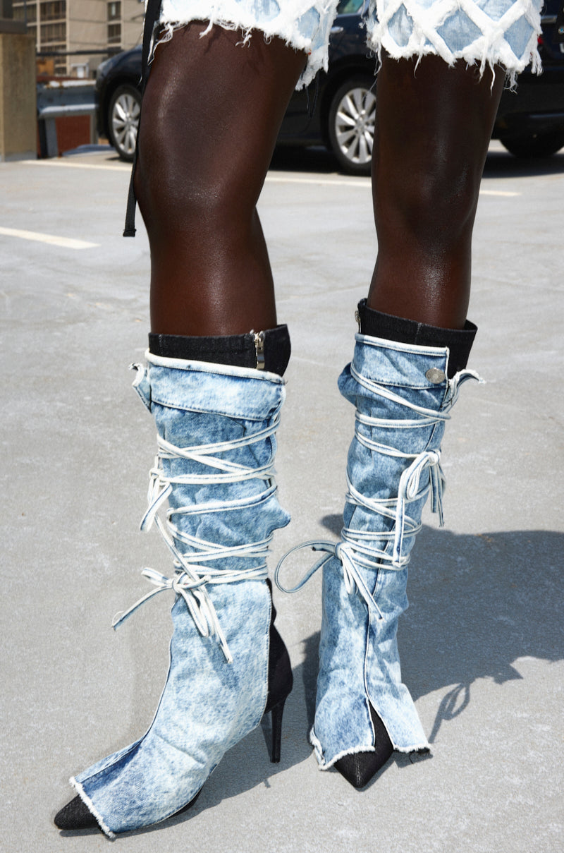 Someone stands on a concrete parking lot wearing the AZALEA WANG VILLAREAL DENIM BOOT WITH TIES—denim knee-high boots with pointed toes, stiletto heels, white laces, detachable denim overlay—paired with frayed denim shorts.