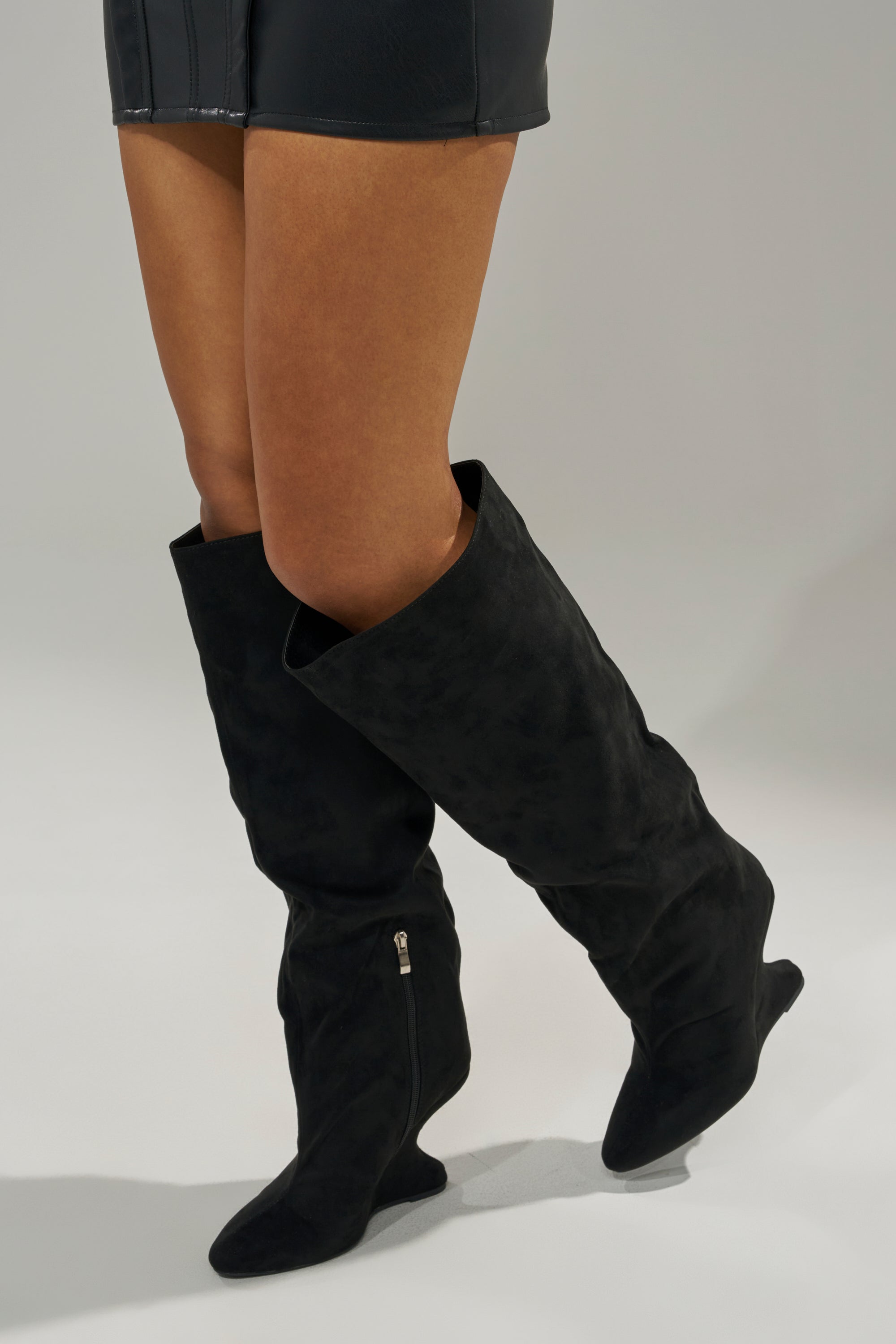 A person wearing a short black skirt and AZALEA WANG VINCE BLACK FAUX SUEDE WEDGE BOOTS with side zippers stands against a neutral background, one foot slightly ahead.