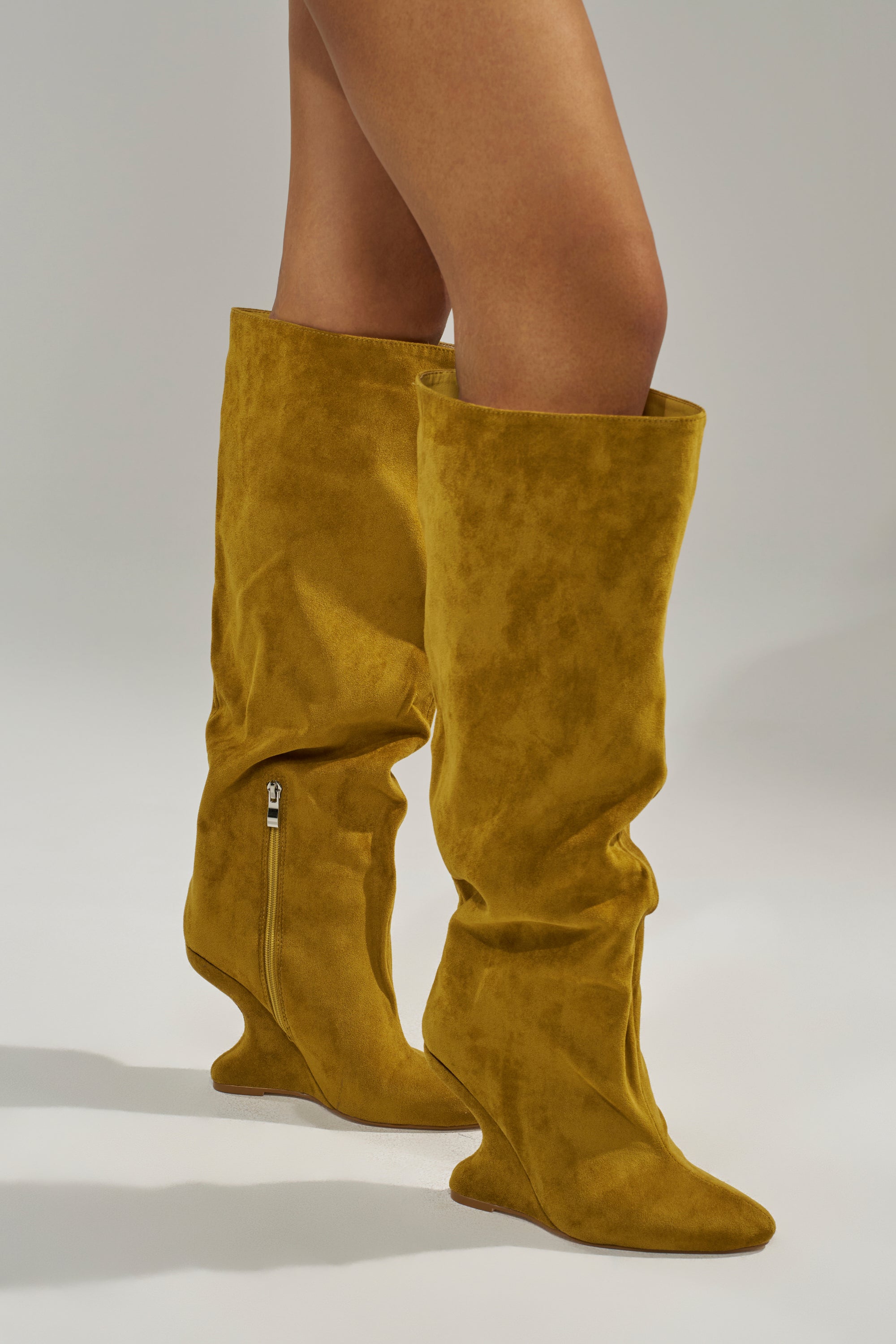 A person models the AZALEA WANG VINCE MUSTARD FAUX SUEDE WEDGE BOOT—pointed toe, knee-high, with a side zipper and slightly slouched design—against a plain light gray background.