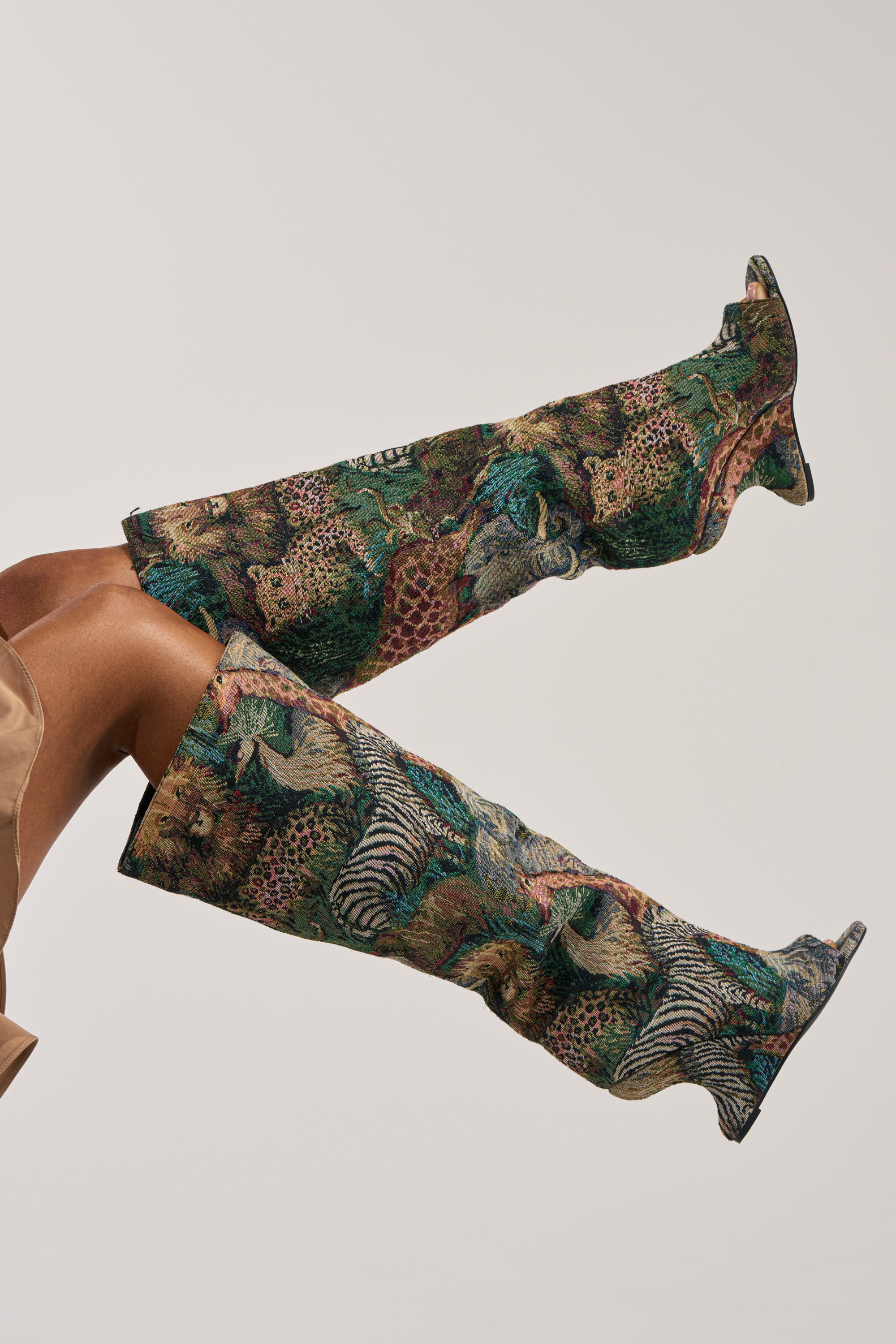 A person with brown skin wears the AZALEA WANG VINCENT ANIMAL WEDGE OPEN TOE BOOT—statement, over-the-knee footwear displaying bold leopard and zebra patterns on a neutral background. Only their legs and part of some beige clothing are visible.