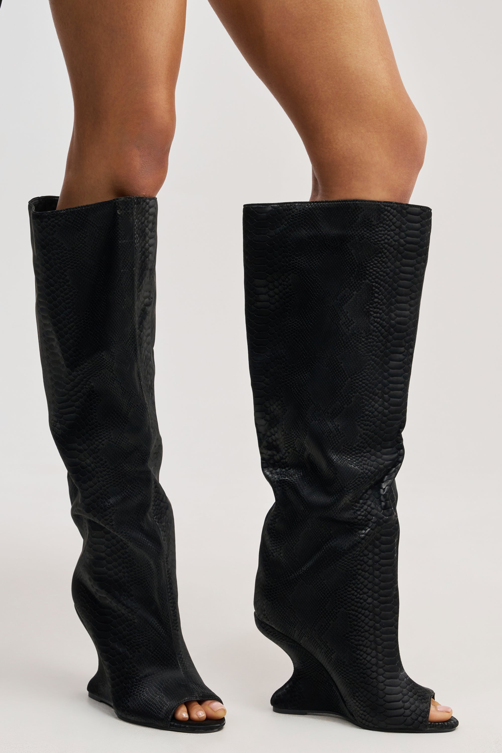 A person models the AZALEA WANG VINCENT BLACK SNAKE OPEN TOE WEDGE BOOT—black snake print, faux leather knee-high boots with open toes and an inverted wedge—against a neutral background.