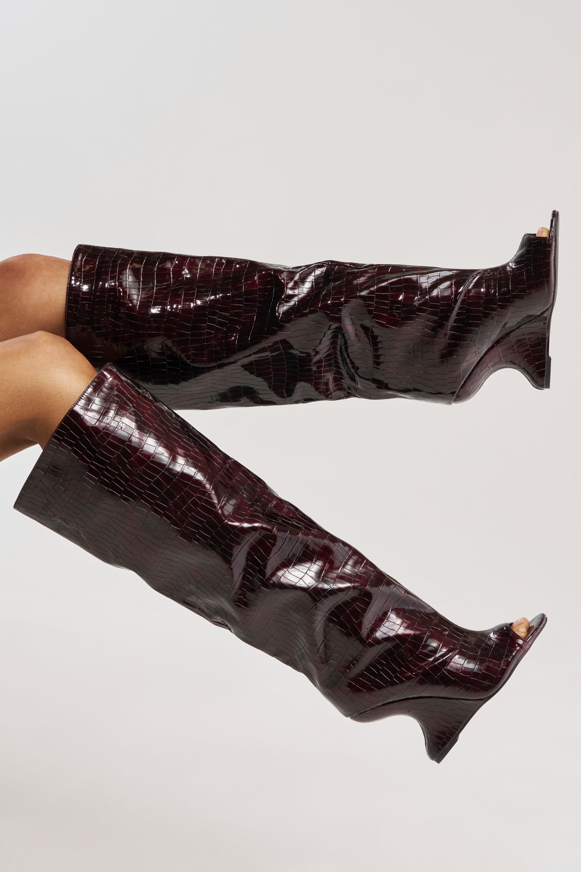 Legs modeled in AZALEA WANG VINCENT BURGUNDY CROC OPEN TOE WEDGE BOOTS, featuring shiny dark burgundy croc embossing and low wedge heels, posed against a plain light background.