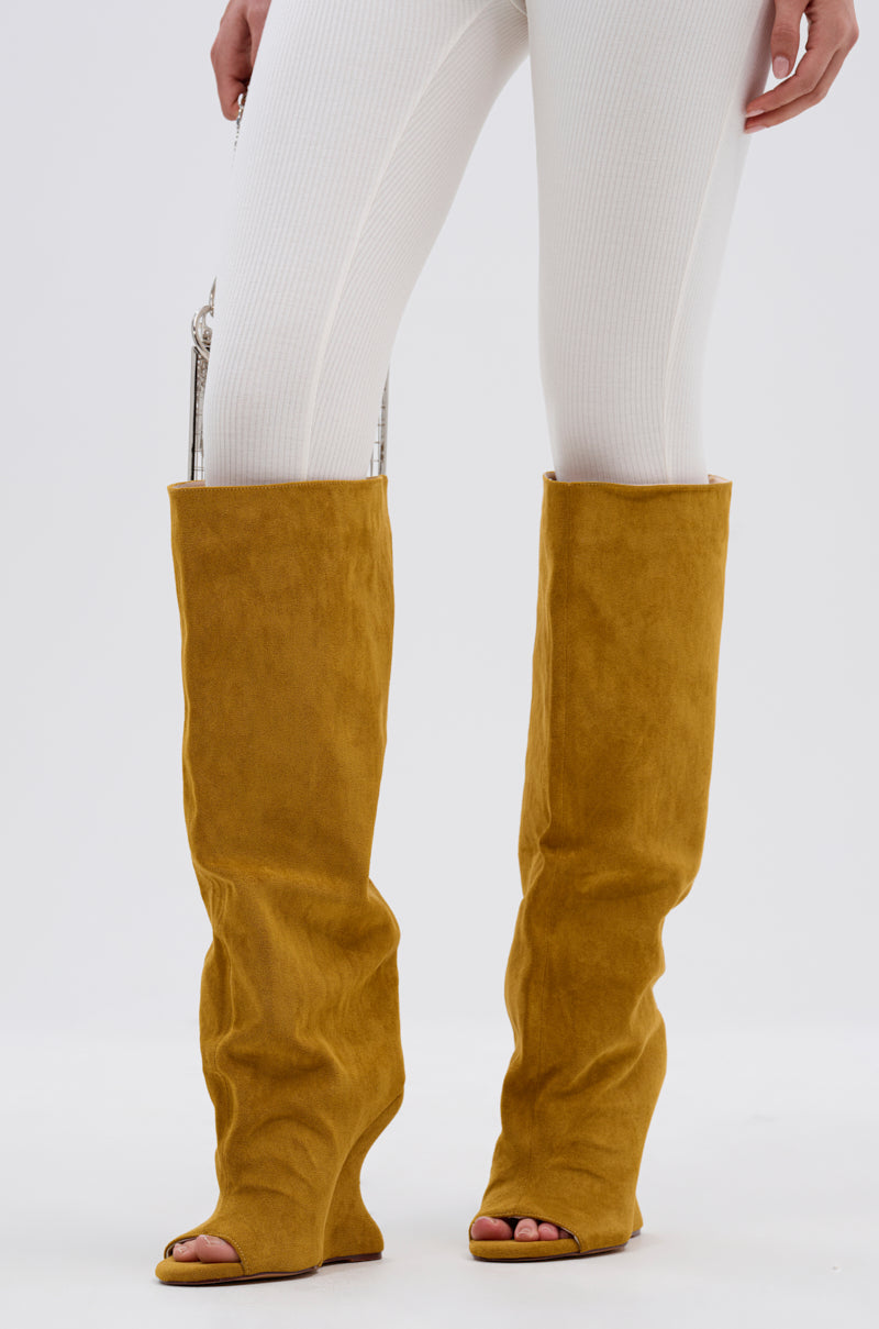 A person wears ribbed white leggings and the AZALEA WANG VINCENT MUSTARD SUEDE OPEN TOE BOOT, featuring an inverted wedge heel and knee-high shaft, standing against a plain white background.