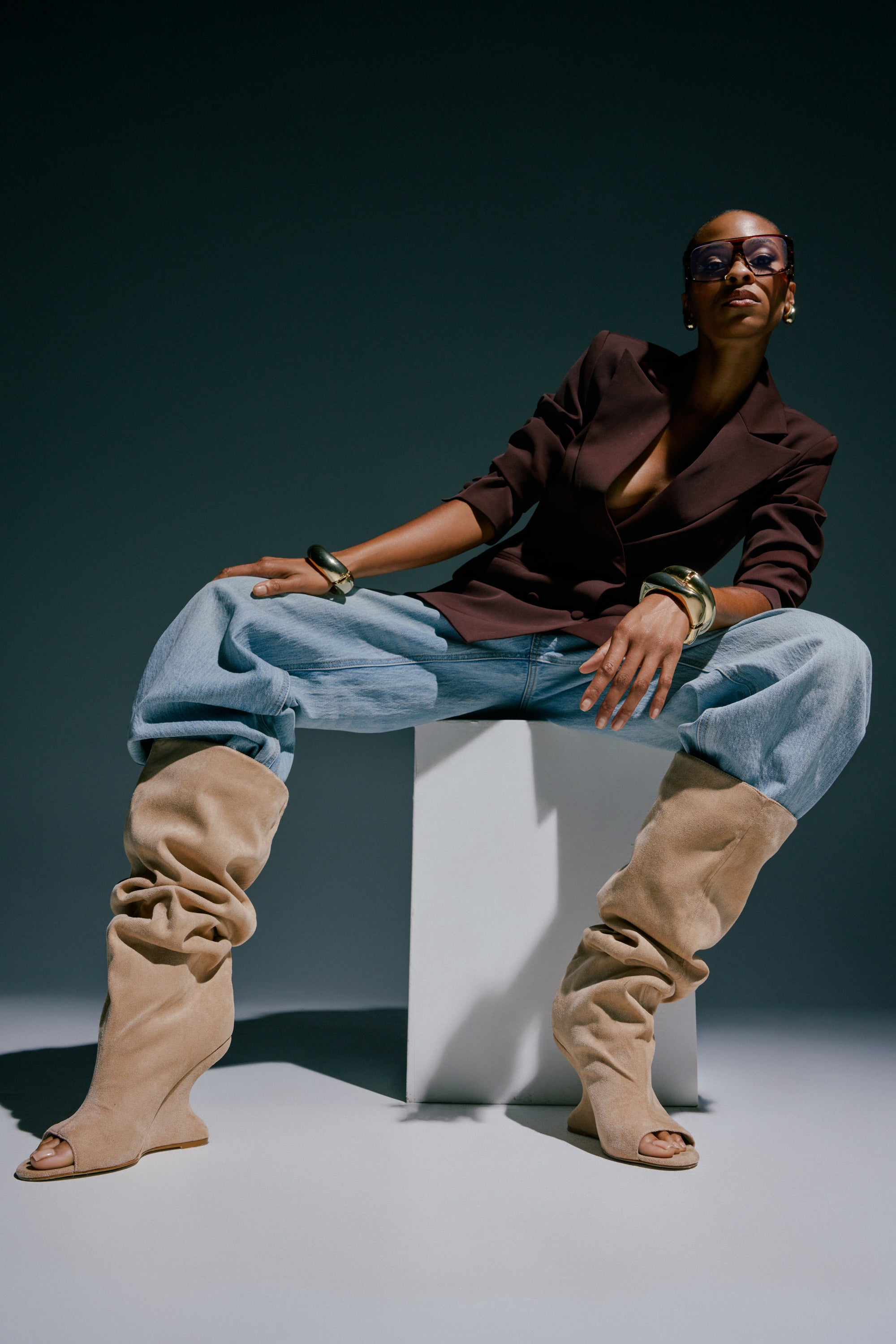 A stylish person poses in sunglasses, a dark blazer, chunky bangles, slouchy light jeans, and the AZALEA WANG VINCENT NUDE FAUX SUEDE OPEN TOE BOOTS while sitting on a white block against a dark background.