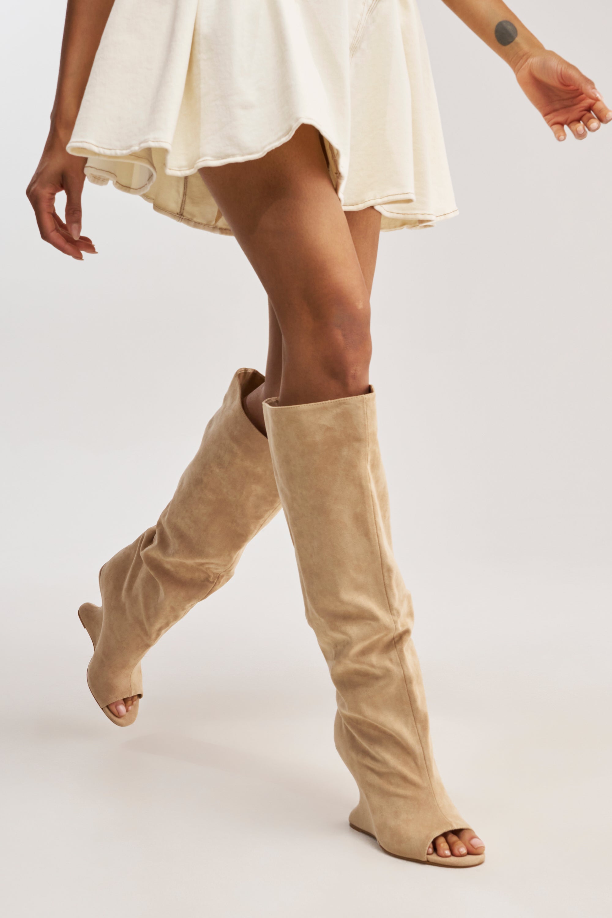 Only the lower half of a person is visible as they walk forward in a short white skirt and AZALEA WANG VINCENT Nude Faux Suede Open Toe Boots, set against a light background.