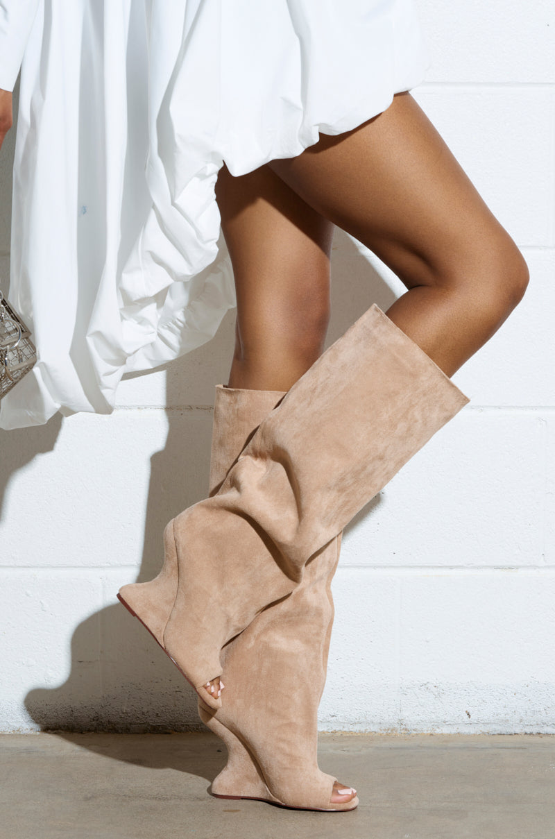 A person in a white dress wears AZALEA WANG VINCENT NUDE OPEN TOE WEDGE BOOTS and stands against a white brick wall, with their left leg crossed over the right as light casts shadows on the floor and wall.