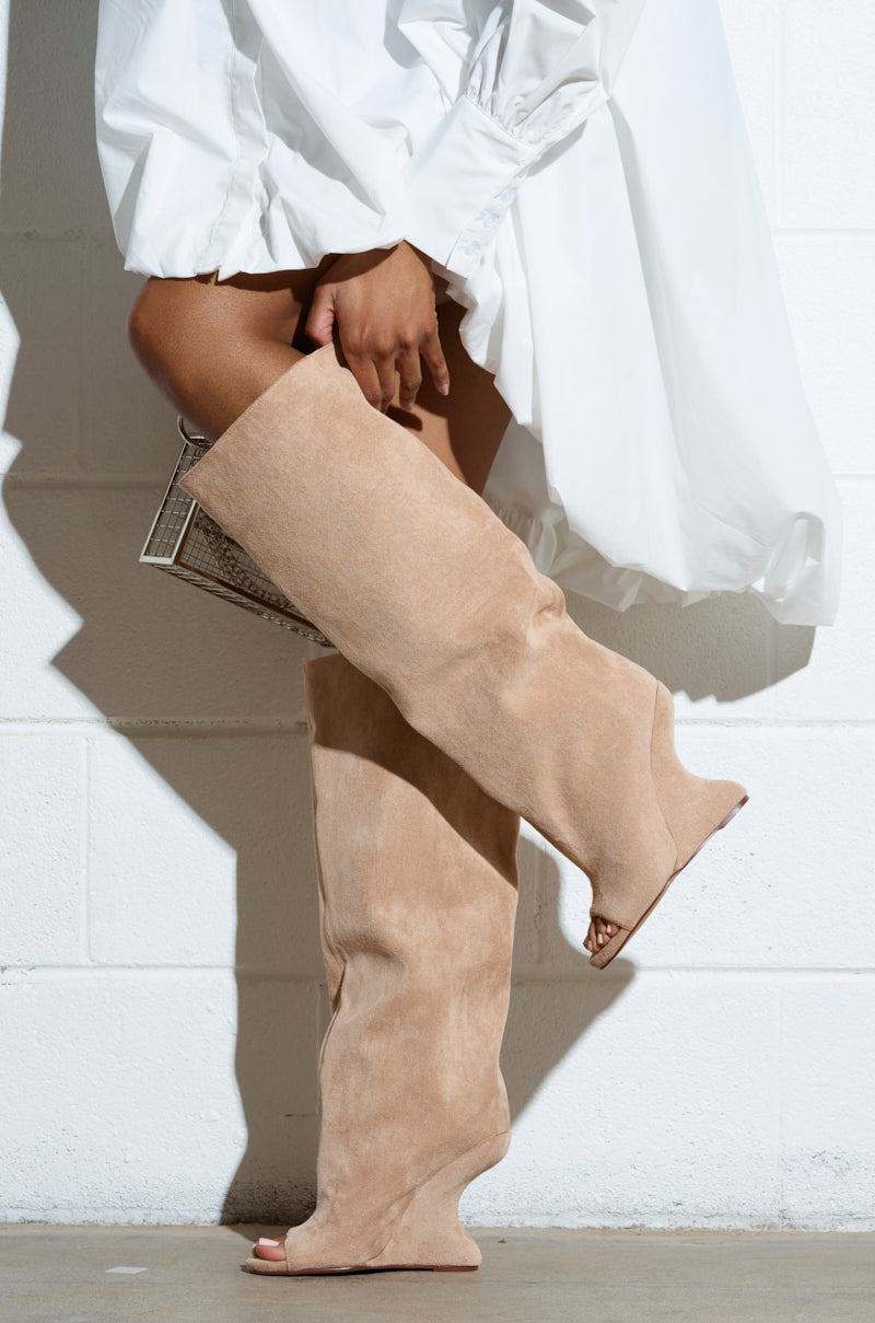 A person in a white dress with puffed sleeves stands against a white brick wall, wearing knee-high AZALEA WANG VINCENT nude open toe wedge boots and holding a small metallic clutch.