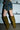 A person stands by a garage door wearing patchwork denim, camouflage shorts, and the AZALEA WANG VINCENT OLIVE FAUX SUEDE OPEN TOE WEDGE BOOT with a metallic accessory on their wrist.