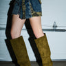 A person stands by a garage door wearing patchwork denim, camouflage shorts, and the AZALEA WANG VINCENT OLIVE FAUX SUEDE OPEN TOE WEDGE BOOT with a metallic accessory on their wrist.