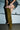 Wearing denim shorts and a vest, a person stands on a concrete floor in front of a closed garage door, pulling up the AZALEA WANG VINCENT OLIVE FAUX SUEDE OPEN TOE WEDGE BOOT over their left leg.