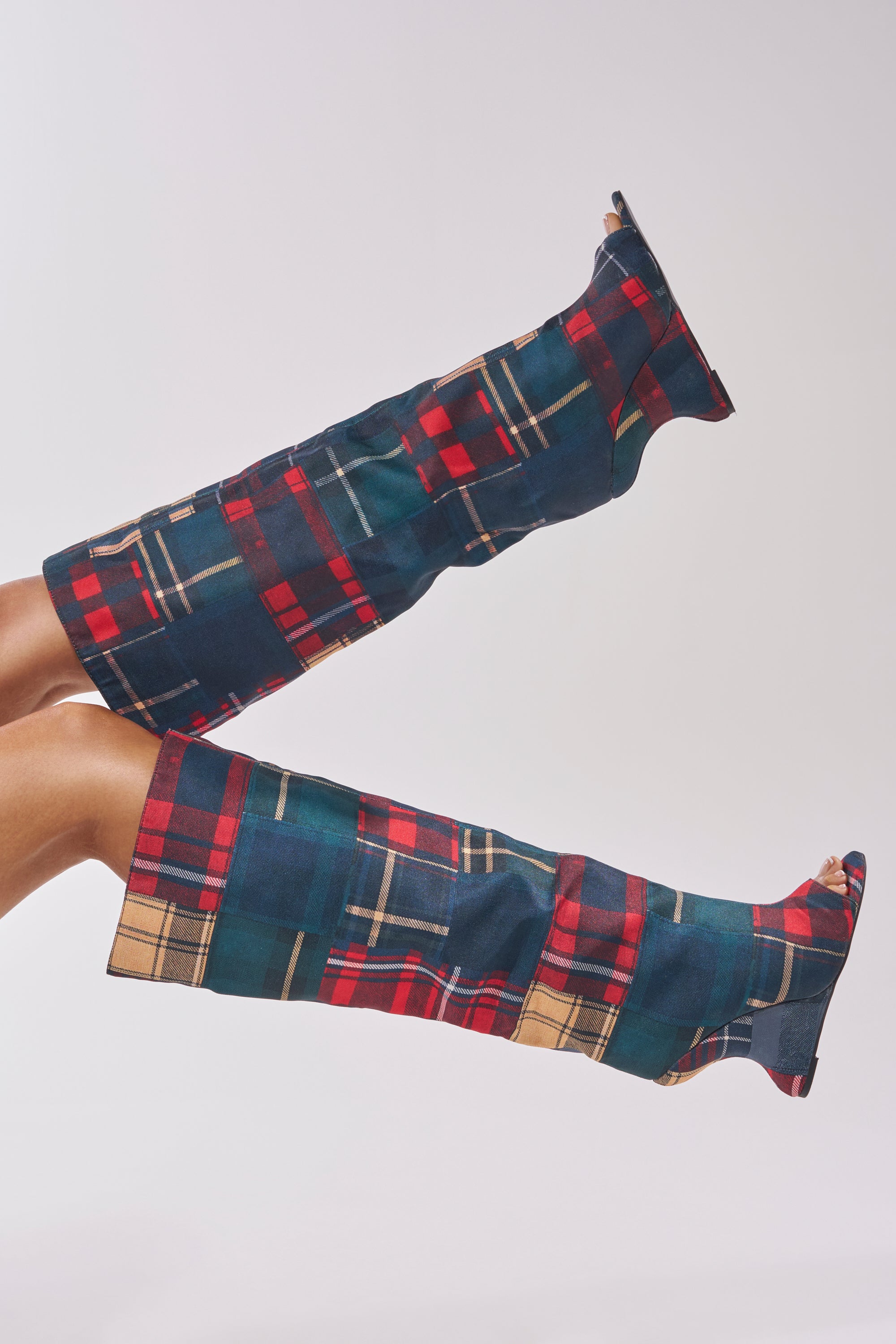 Two legs wearing the AZALEA WANG VINCENT PLAID PRINTED OPEN TOE BOOT—tall, over-the-knee boots with a vibrant red, green, yellow, and blue plaid pattern—are posed horizontally against a light background.