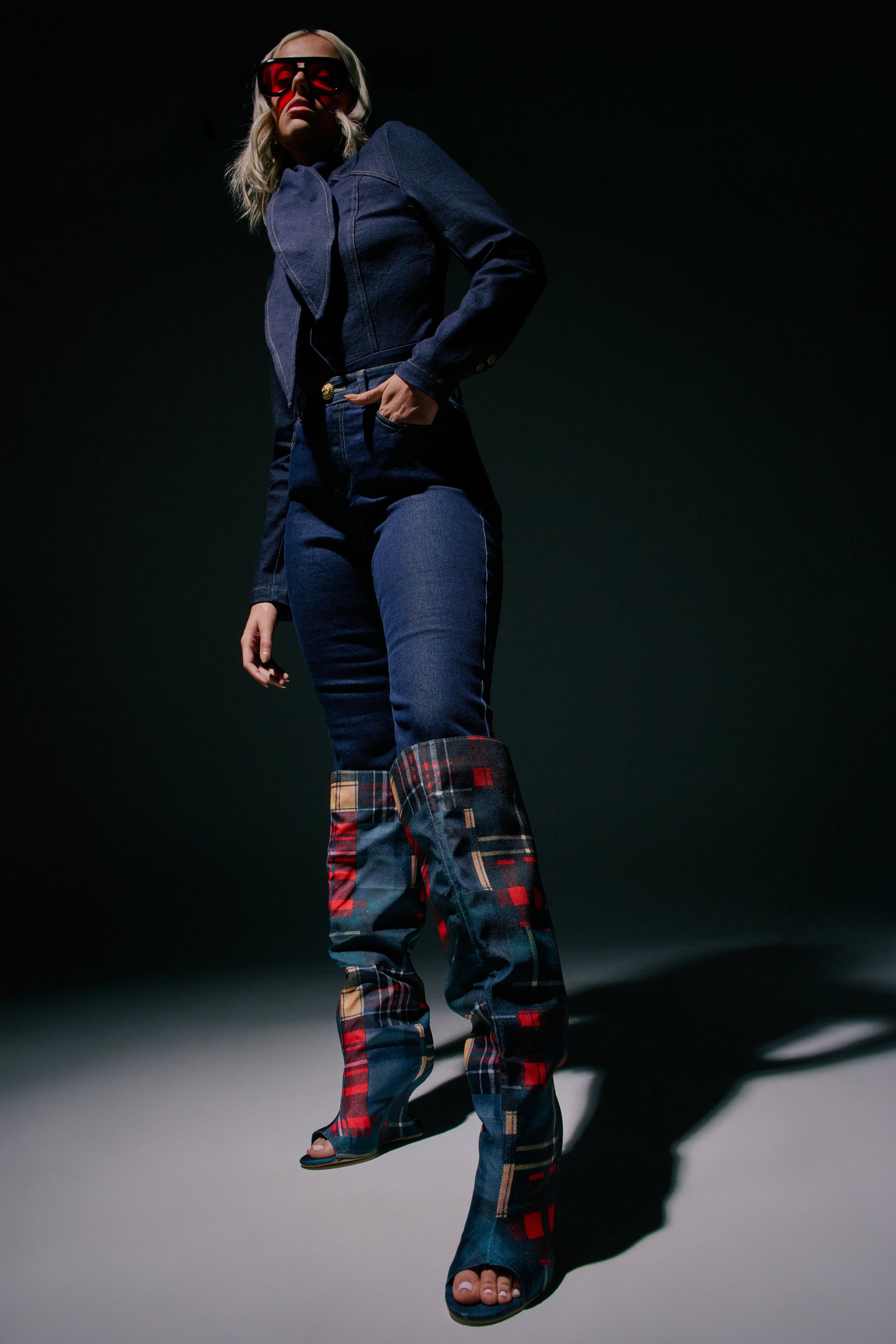 A woman stands confidently against a dark background, casting a shadow, dressed in a dark denim jacket and jeans, red sunglasses, and striking AZALEA WANG VINCENT PLAID PRINTED OPEN TOE BOOTS featuring an inverted wedge heel.